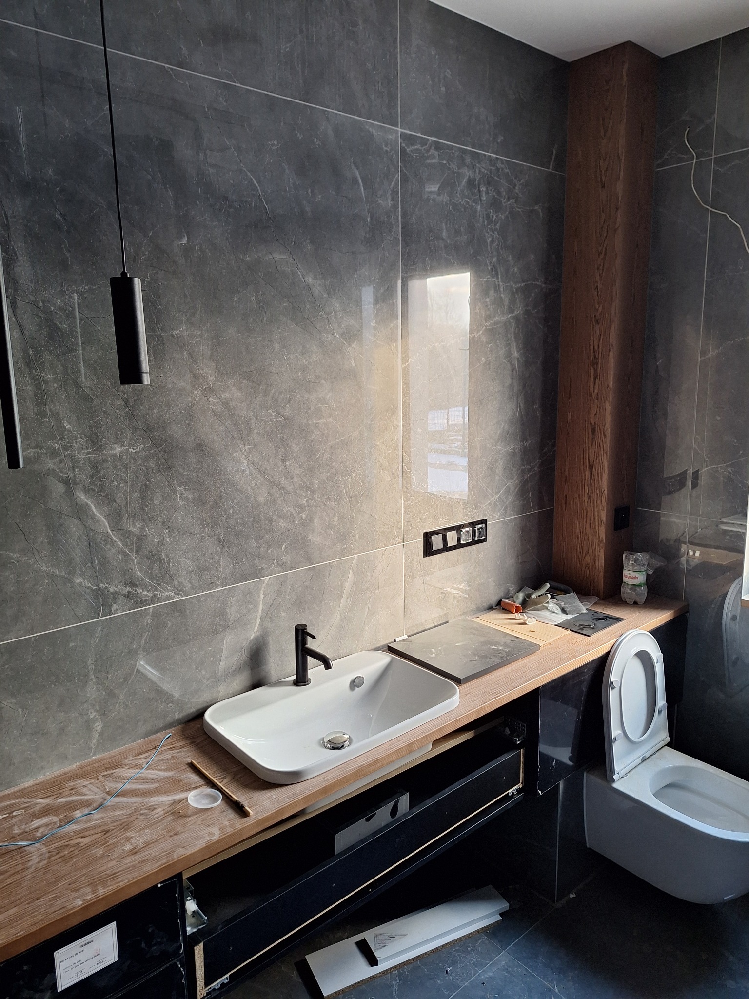 Wnętrze łazienki w trakcie remontu, widoczne szare płytki imitujące marmur na ścianach, biała umywalka z czarną baterią, toaleta oraz niedokończona zabudowa szafkowa z drewnianym blatem.