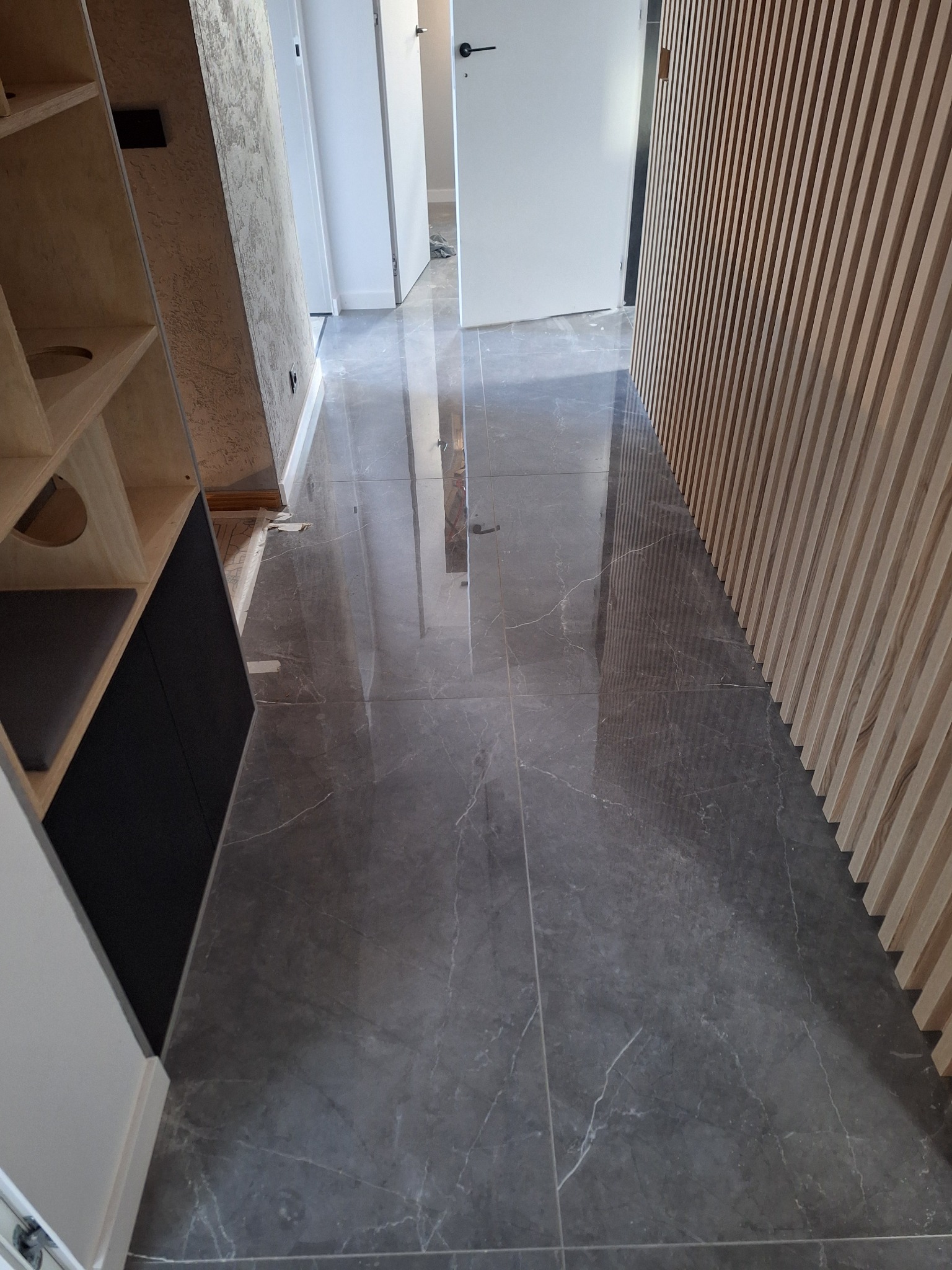 Szara marmurowa podłoga z połyskiem w nowo wykończonym korytarzu, widoczne drewniane lamele ścienne i fragmenty zabudowy meblowej.