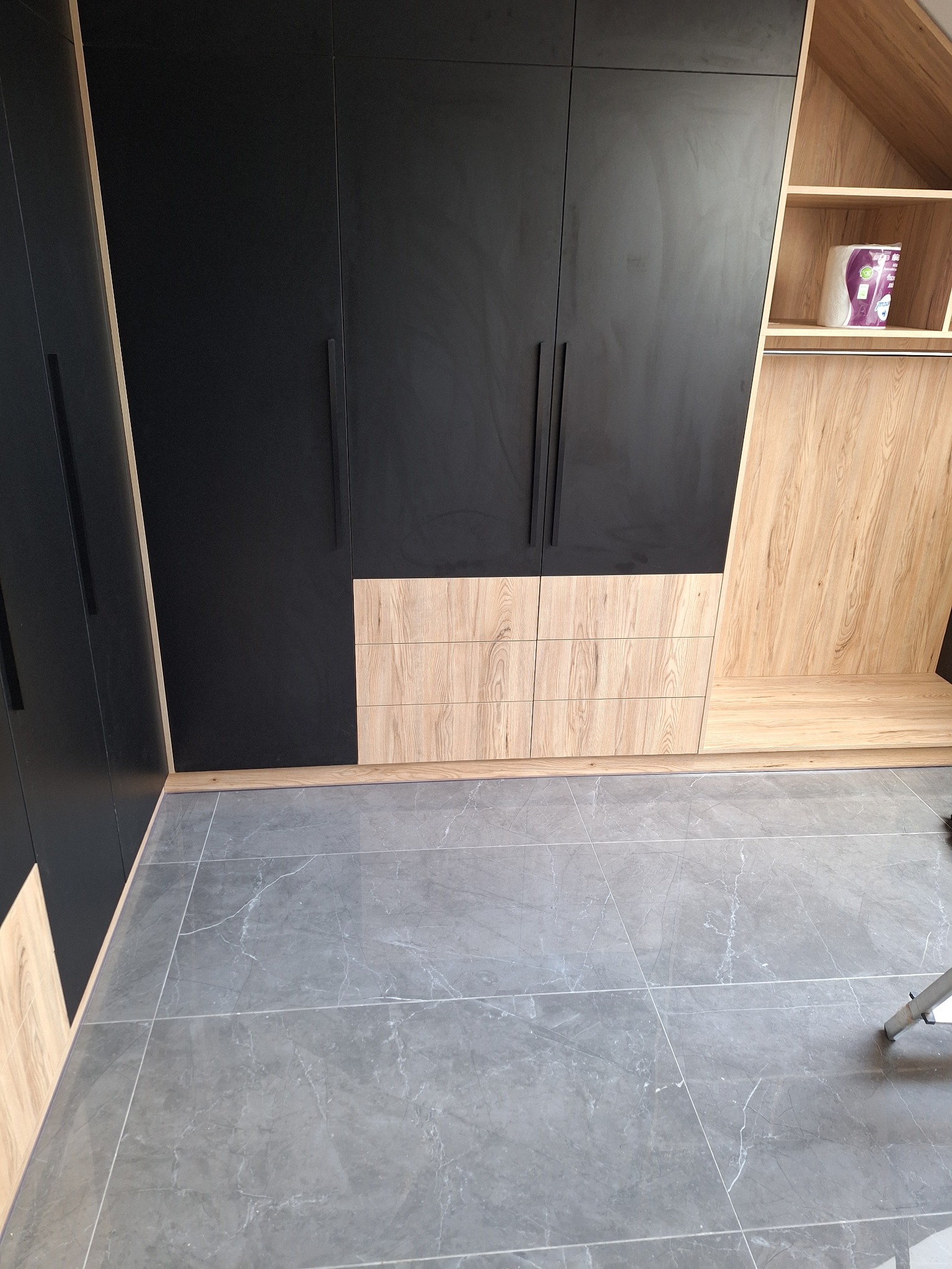 Czarno-drewniana zabudowa garderoby z szufladami i półkami, widok na szare płytki podłogowe.