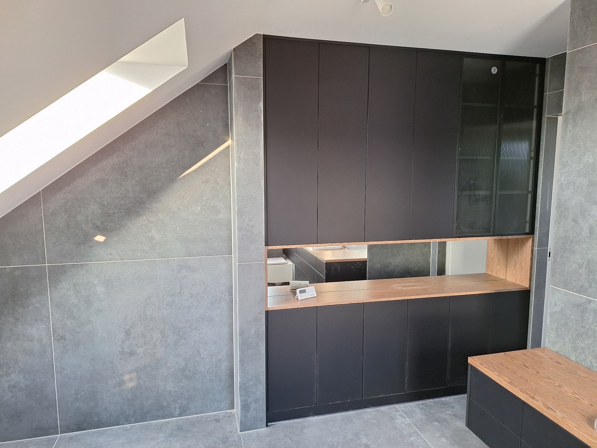 Szara łazienka na poddaszu z zabudową szafy z czarnymi frontami i drewnianymi półkami, widoczne szare płytki na ścianach i podłodze, oraz okno dachowe.