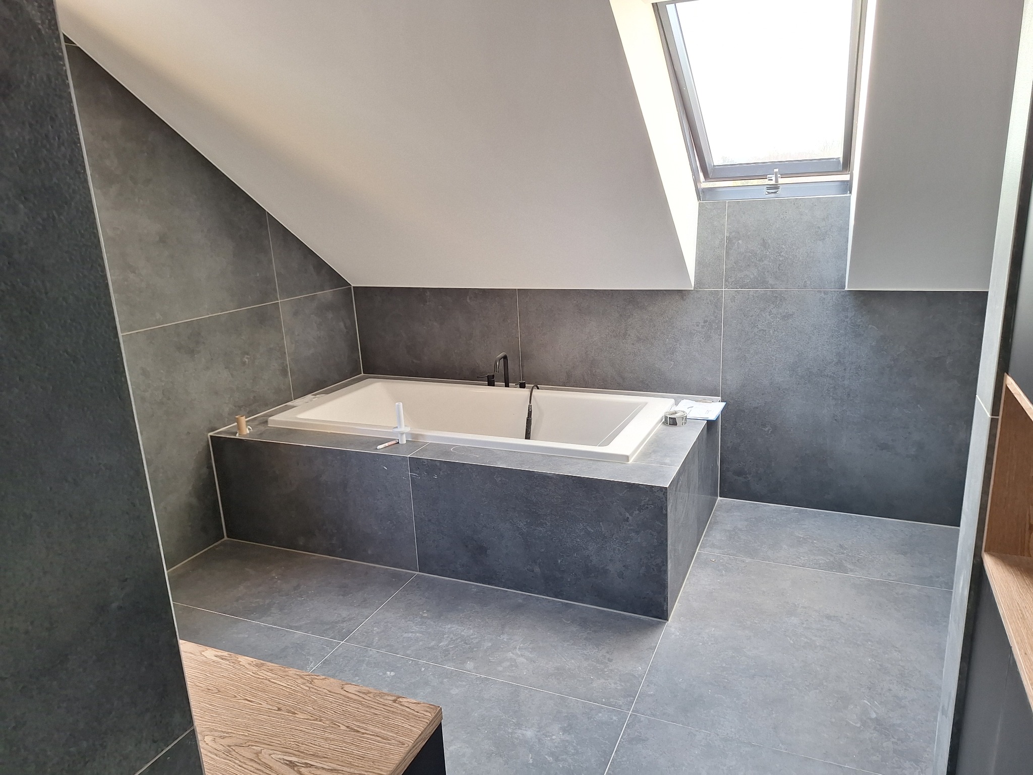 Nowoczesna łazienka na poddaszu z wanną obudowaną ciemnoszarymi płytkami, podłoga i ściany wyłożone płytkami w podobnym odcieniu, okno dachowe wpuszczające naturalne światło, widoczne akcesoria...
