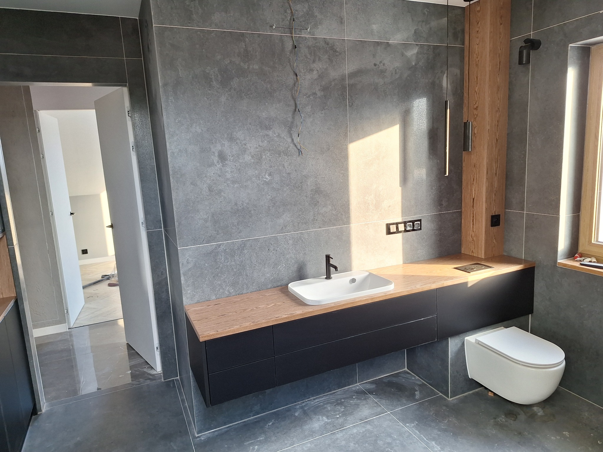 Nowoczesna łazienka z szarymi, wielkoformatowymi płytkami, drewnianym blatem pod umywalką i wiszącą toaletą; widoczne minimalistyczne oświetlenie i otwarte drzwi do innego pomieszczenia.