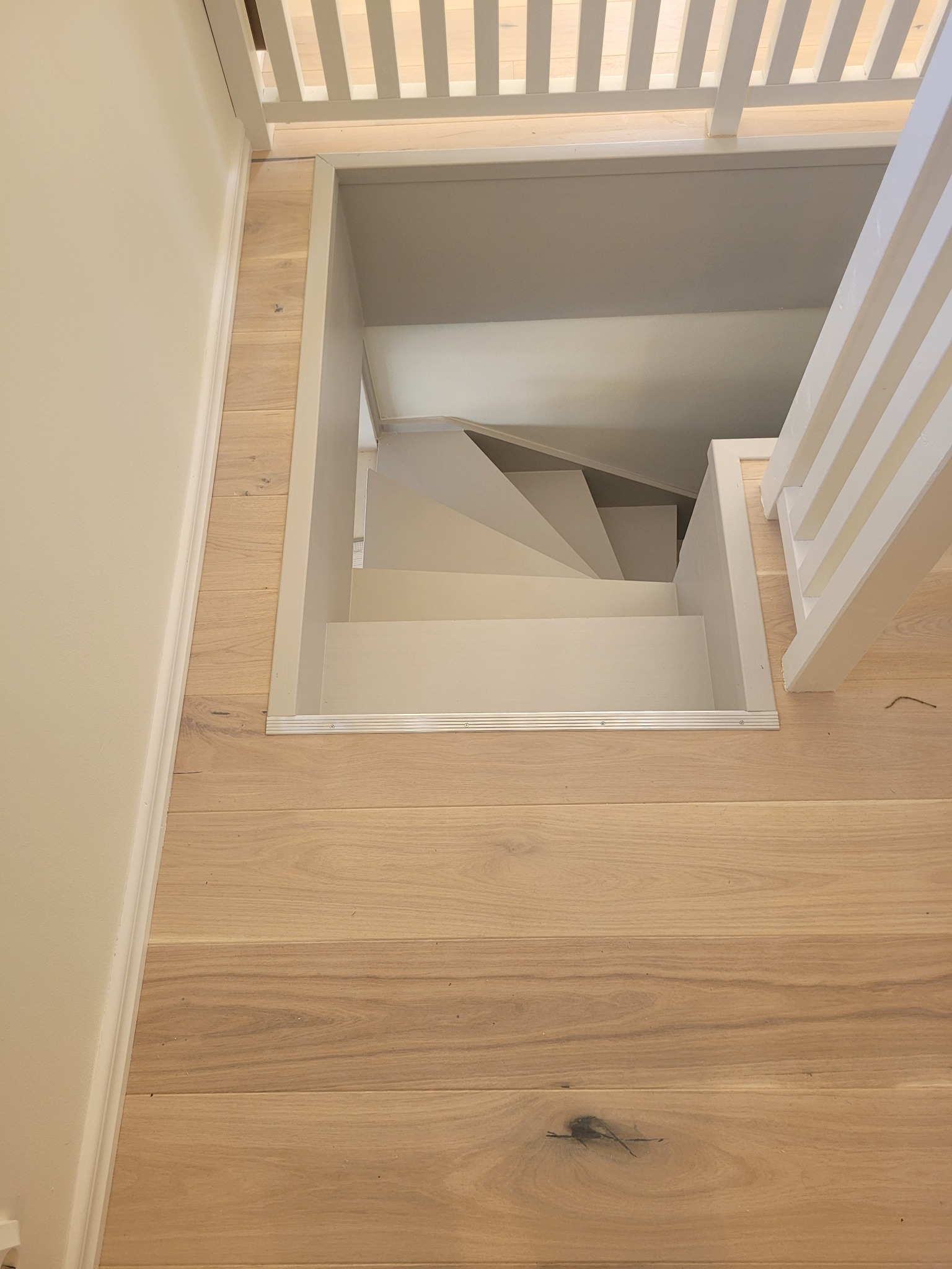 Widok z góry na pomalowane na biało schody prowadzące w dół, otoczone drewnianą podłogą i białą balustradą.