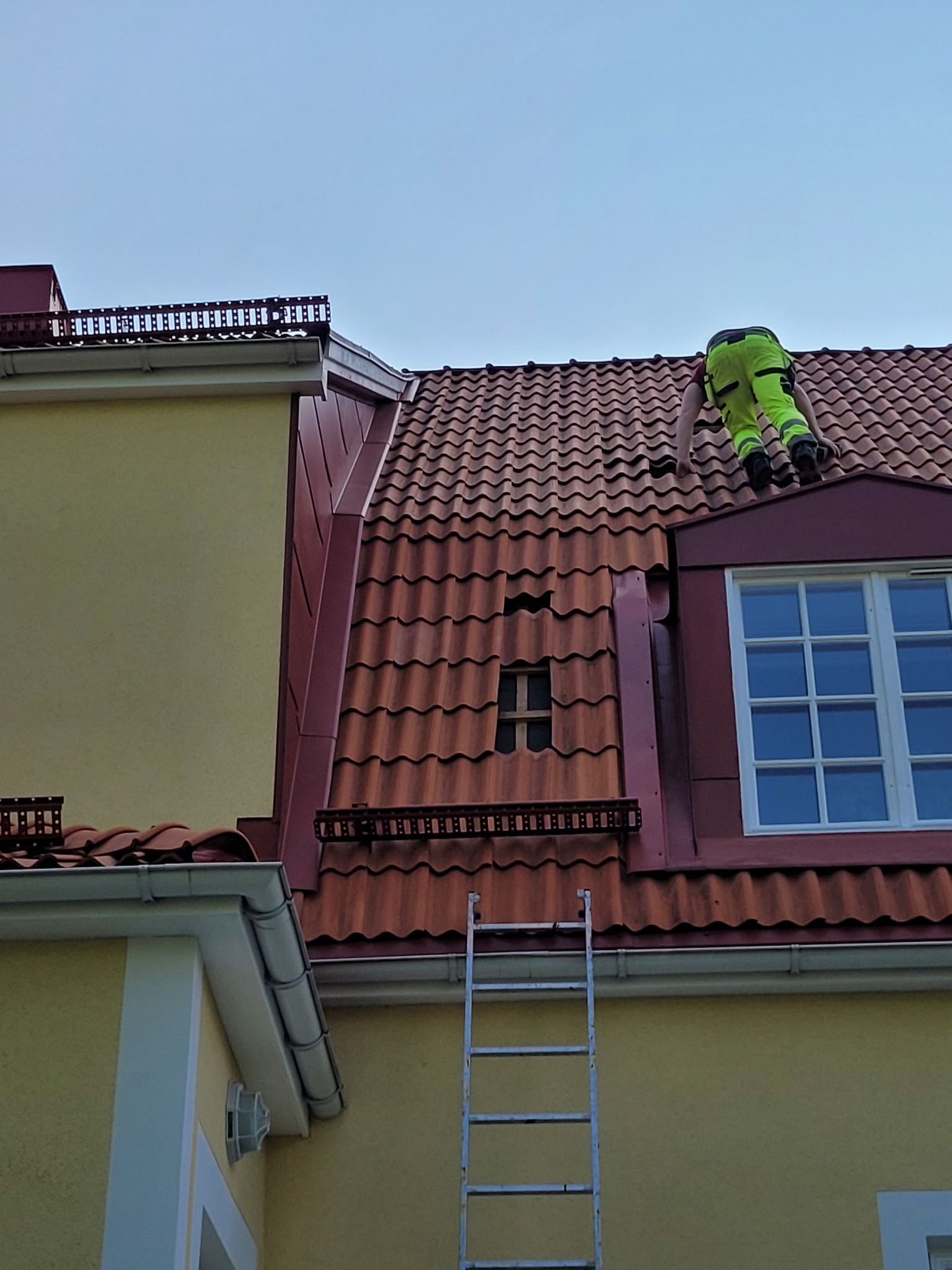 Pracownik w neonowo-zielonym kombinezonie na dachu z czerwonej dachówki, obok okna dachowego i drabiny, w tle błękitne niebo.