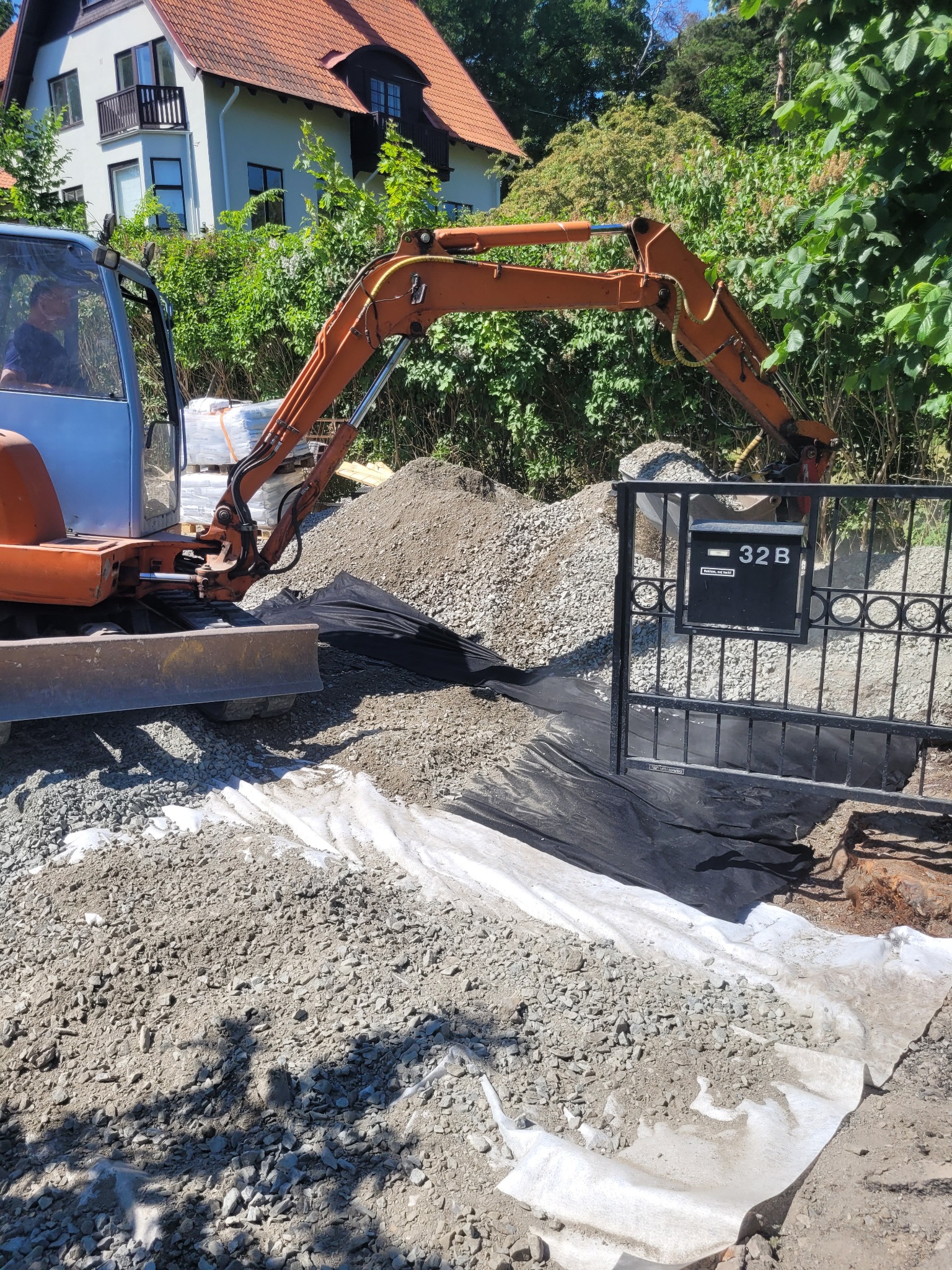 Pomarańczowa koparka na posesji przed domem, przygotowująca teren pod budowę lub remont, widoczna czarna geowłóknina i biała agrowłóknina, oraz brama z numerem 32B.
