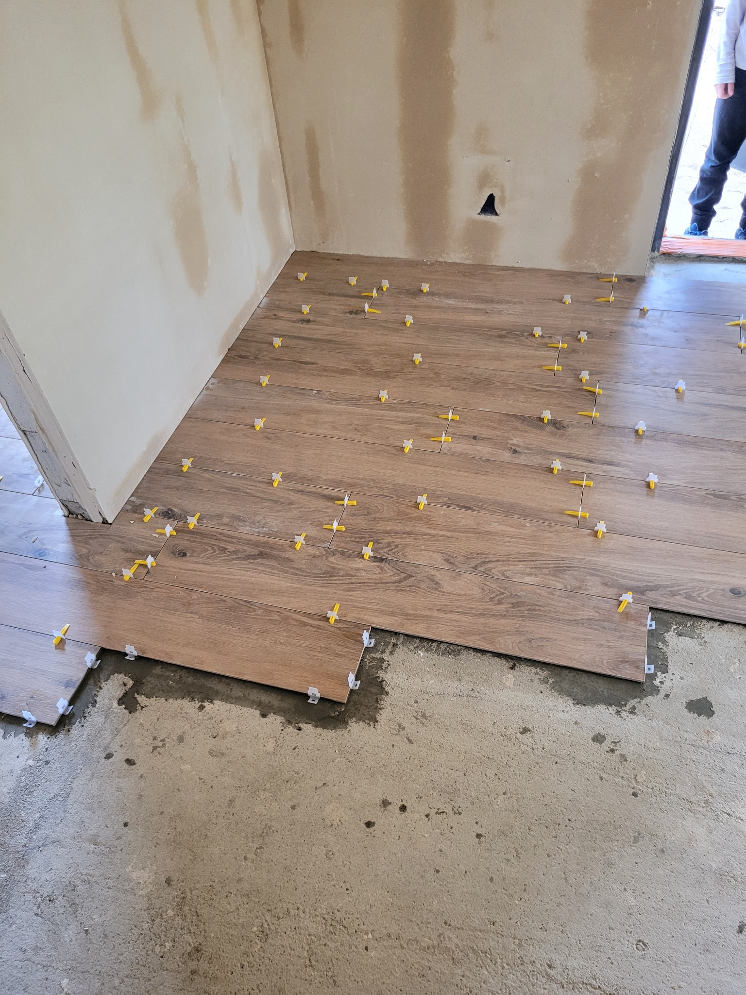 Układanie brązowych płytek imitujących drewno na podłodze, z użyciem żółtych krzyżyków dystansowych, widoczny fragment surowej ściany.