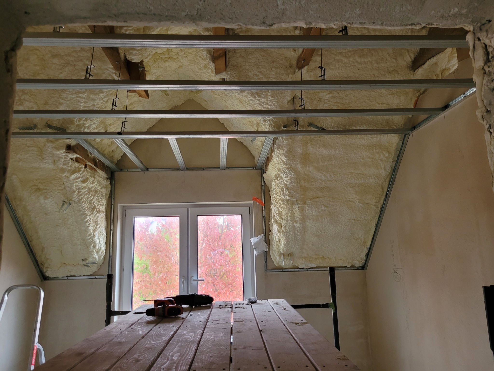 Poddasze w trakcie ocieplania pianą PUR, widoczne profile metalowe pod konstrukcję sufitu, narzędzia na drewnianym stole roboczym, okno z widokiem na jesienne drzewa.