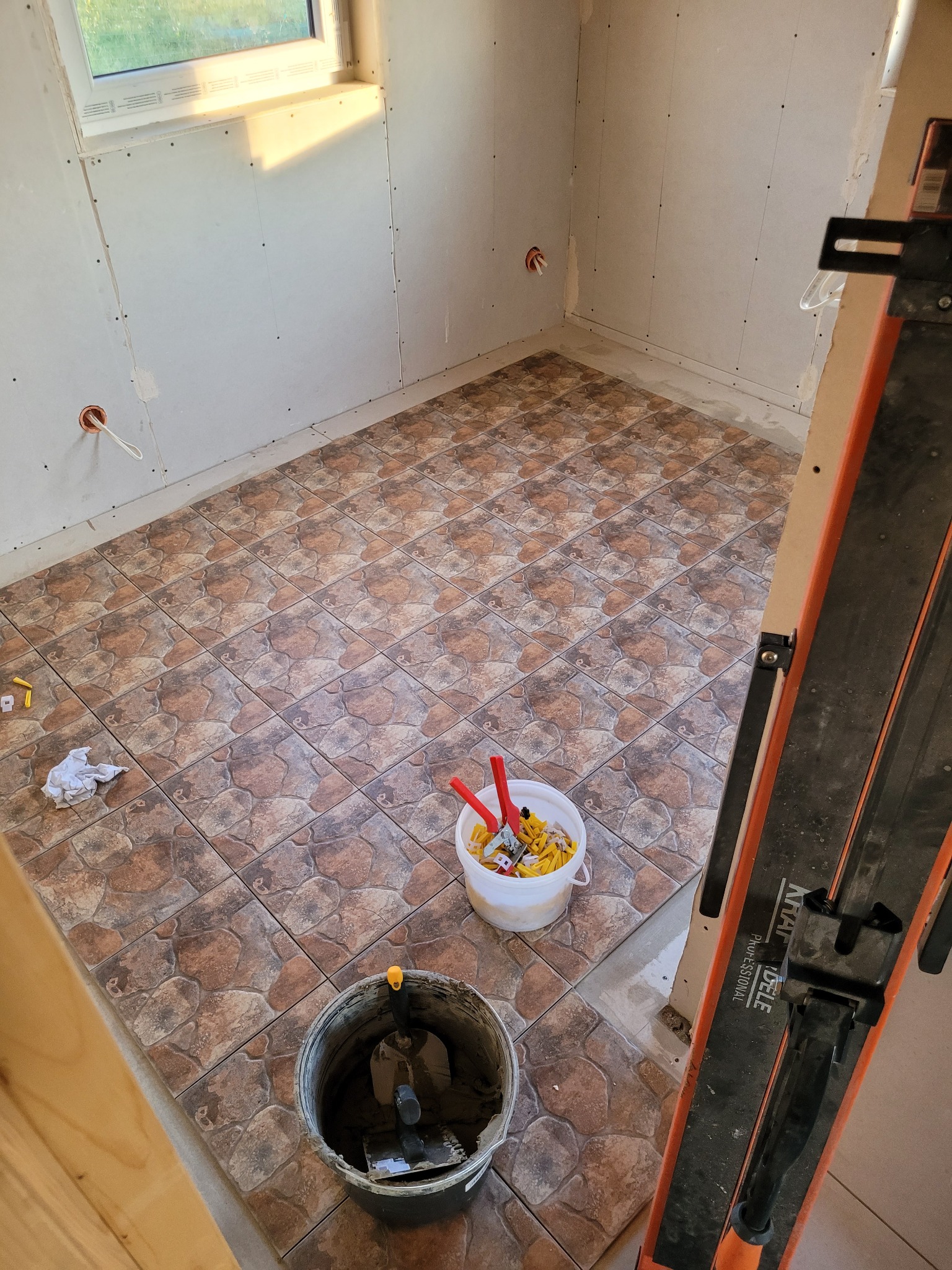 Wnętrze w trakcie prac wykończeniowych: brązowe płytki podłogowe o wzorze kamienia, ściany wykończone płytami, widoczny sprzęt do cięcia płytek, wiadro z zaprawą i narzędziami glazurniczymi,...