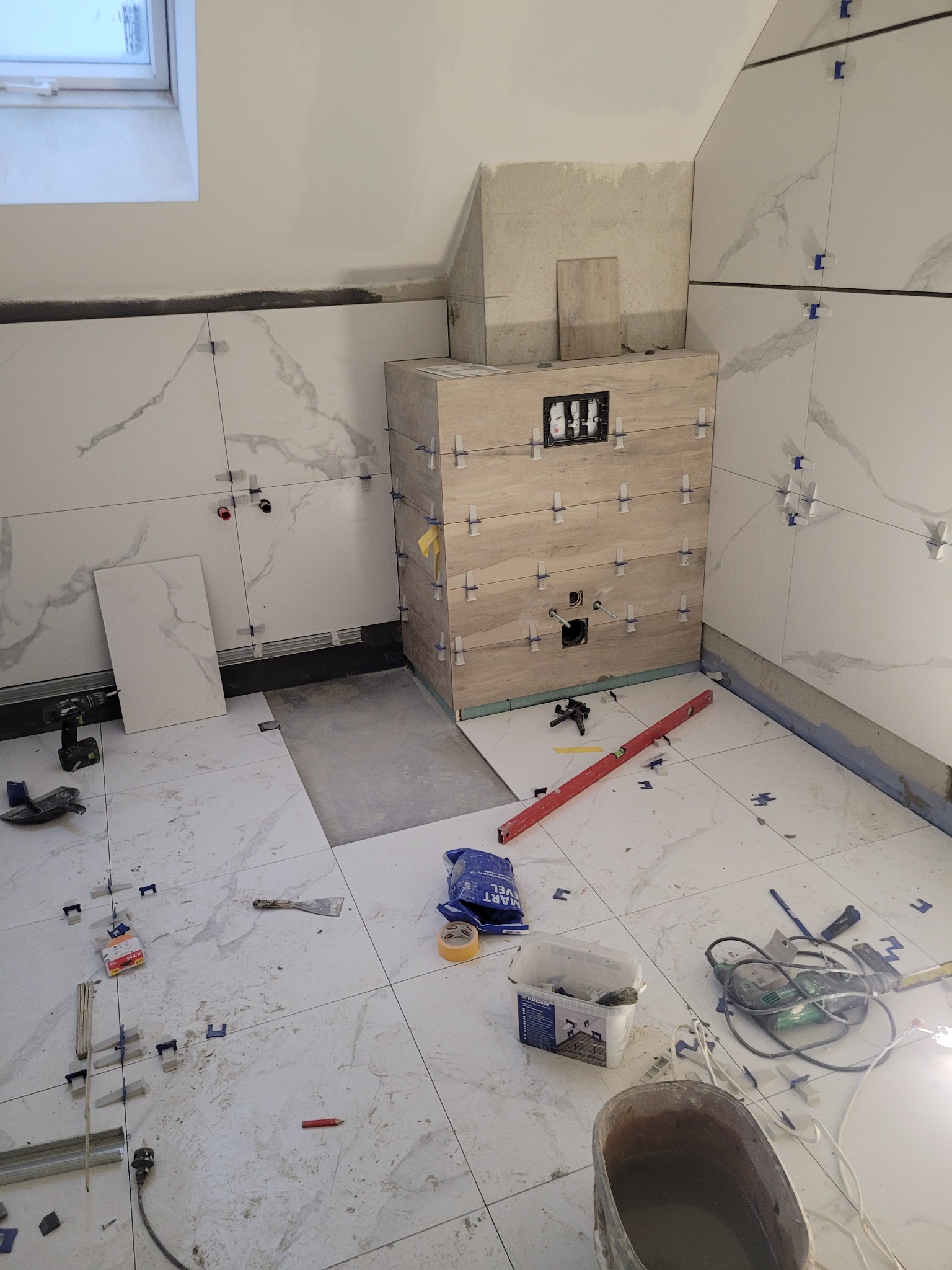 Montaż płytek imitujących marmur w łazience na poddaszu, widoczne narzędzia, poziomica, krzyżyki dystansowe i szafka pod umywalkę w trakcie obkładania płytkami.