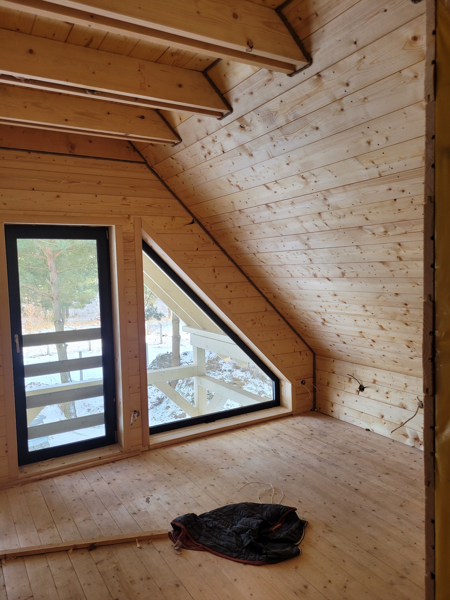 Wnętrze drewnianego domu z ukośnym sufitem i oknem trójkątnym, widoczne drewniane ściany, podłoga, czarna kurtka leżąca na podłodze, częściowo widoczne instalacje elektryczne.