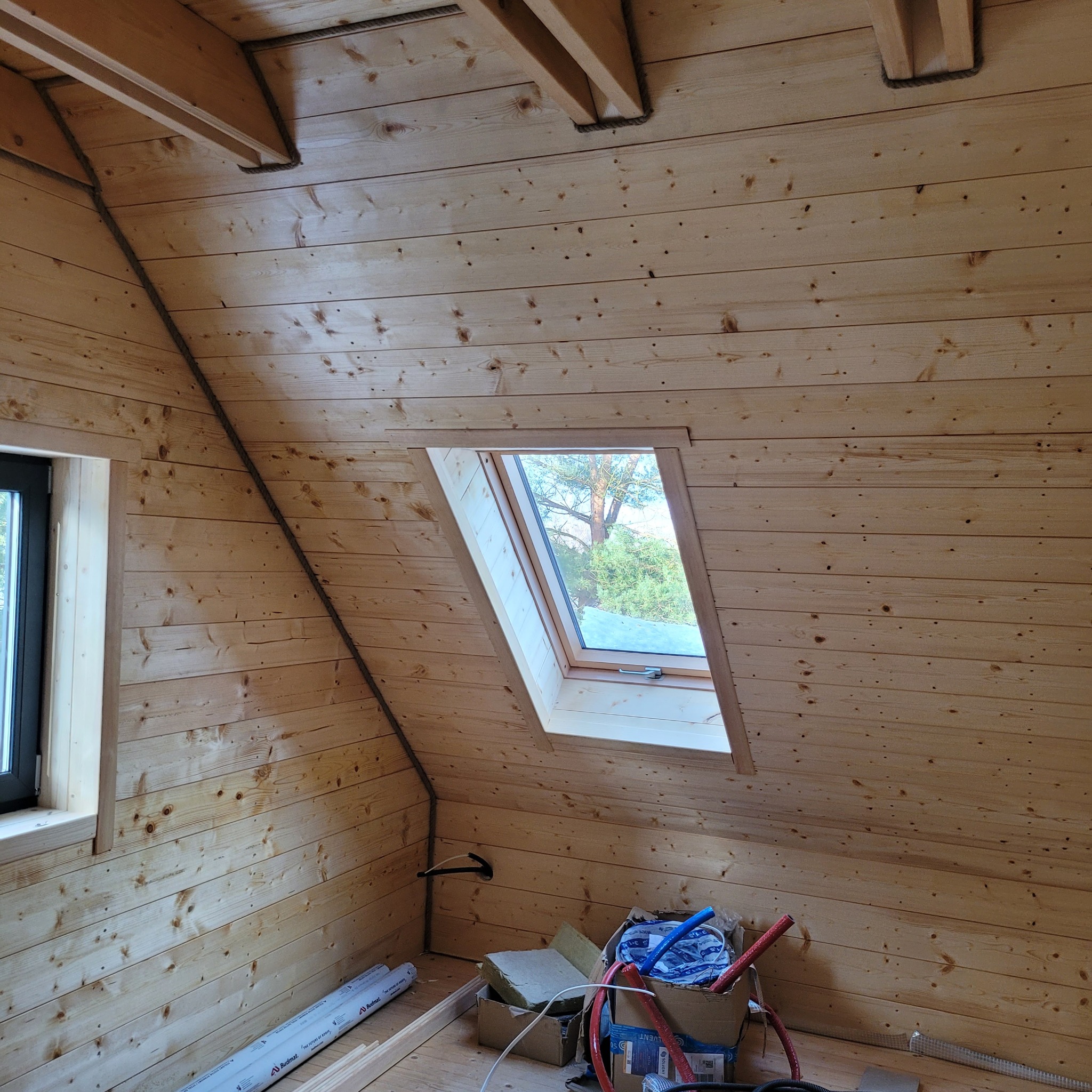 Drewniane wykończenie poddasza z oknem dachowym w Drawsku Pomorskim, widoczne belki stropowe z dekoracyjnym sznurem, częściowo wykończona ściana z boazerii, na podłodze materiały budowlane...