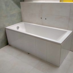 MORIS SOLUTIONS SPÓŁKA Z OGRANICZONĄ ODPOWIEDZIALNOŚCIĄ - Biała wanna obudowana płytkami, z widocznym poziomem na półce i szarymi płytkami w tle, po remoncie łazienki.