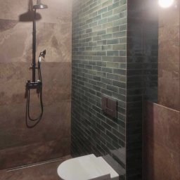 MORIS SOLUTIONS SPÓŁKA Z OGRANICZONĄ ODPOWIEDZIALNOŚCIĄ - Wykończona łazienka z brązowymi płytkami imitującymi kamień i ścianą z zielonej, cegiełkowej glazury, widoczny czarny zestaw prysznicowy retro i podwieszana toaleta.