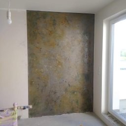 MORIS SOLUTIONS SPÓŁKA Z OGRANICZONĄ ODPOWIEDZIALNOŚCIĄ - Fragment ściany z dekoracyjną, wielobarwną fakturą przypominającą naturalny kamień, obok okno i widoczne narzędzia malarskie oraz wiadro w pomieszczeniu w trakcie prac wykończeniowych.