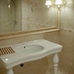 MORIS SOLUTIONS SPÓŁKA Z OGRANICZONĄ ODPOWIEDZIALNOŚCIĄ - Jasna łazienka w Raniżowie z beżowymi płytkami, lustrem w ozdobnej ramie odbijającym kabinę prysznicową i umywalką na toczonych nogach z drewnianą kratką ociekową.