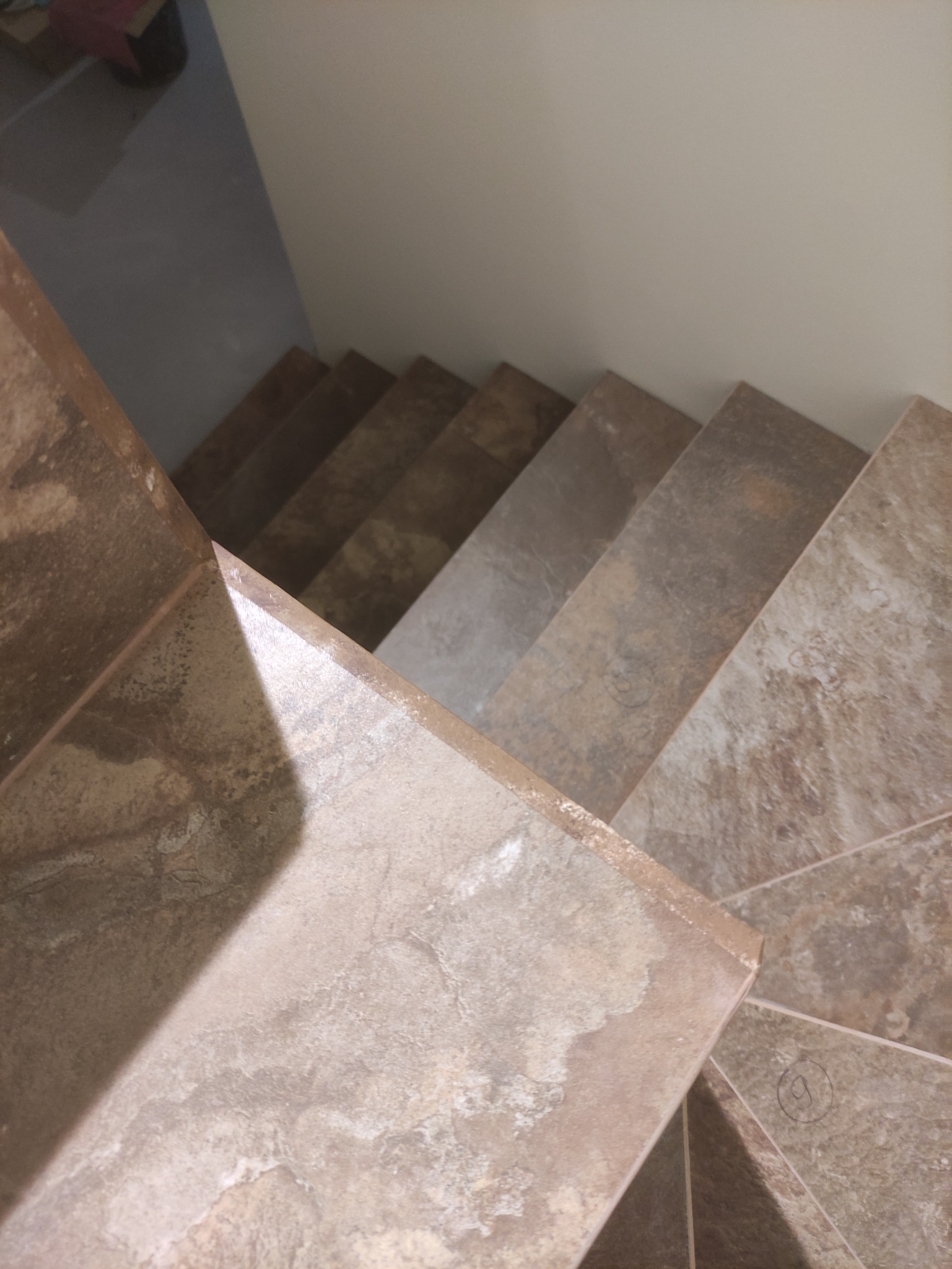 Schody wyłożone brązowymi płytkami imitującymi kamień, widok z góry, z widocznym załamaniem biegu schodów i fragmentem jasnej ściany.