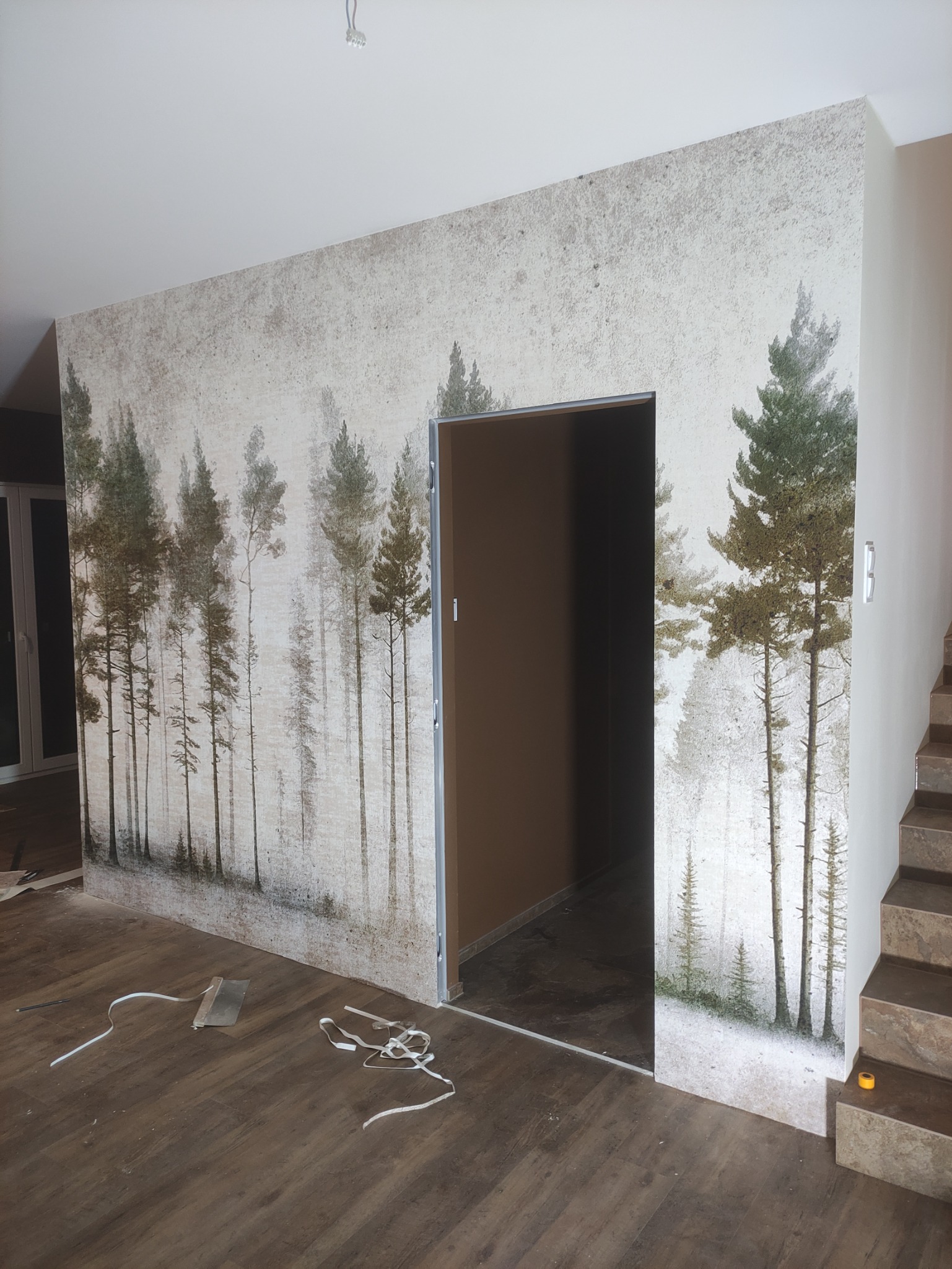 Ściana w pokoju z fototapetą przedstawiającą las, widoczny fragment podłogi z panelami, otwór drzwiowy z brązowymi drzwiami oraz fragment schodów z brązowymi płytkami.