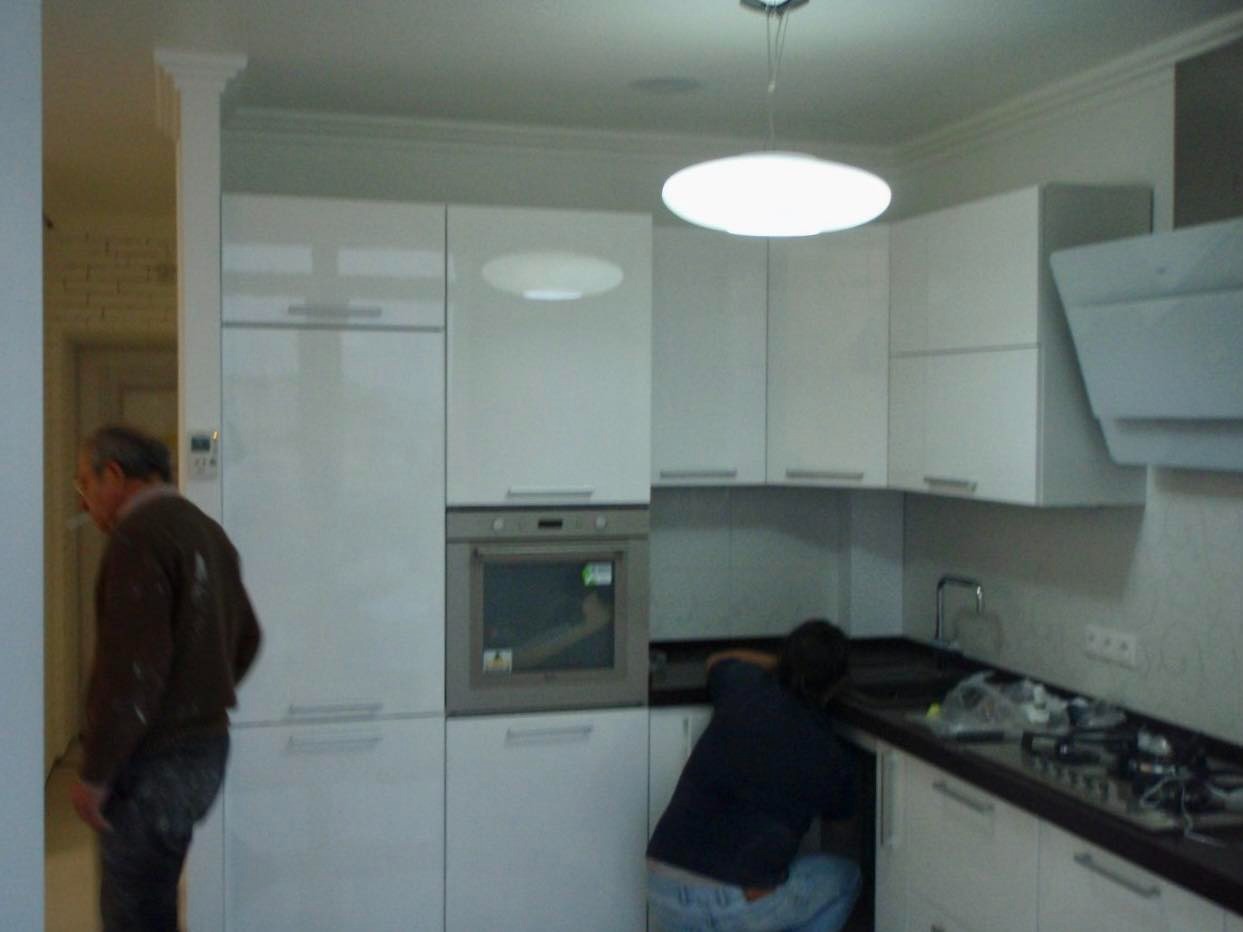 Montaż nowej, białej zabudowy kuchennej z czarnym blatem, widoczny pracownik instalujący zlew i palnik gazowy, oraz drugi pracownik z papierosem w ustach.