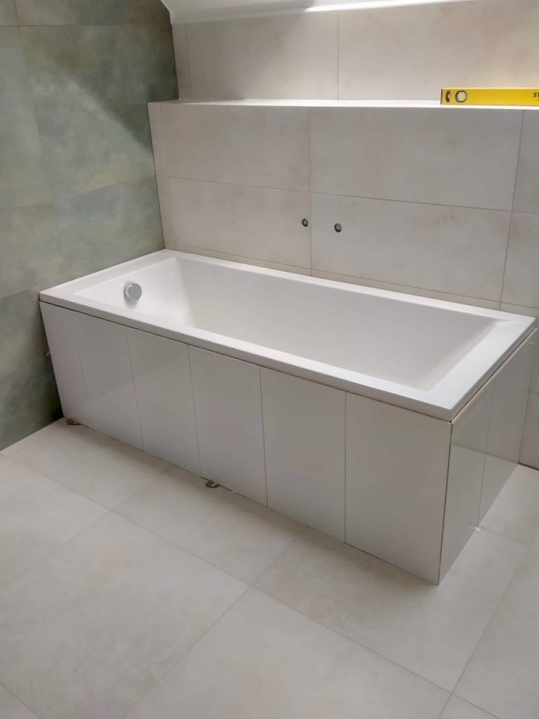 Biała wanna obudowana płytkami, z widocznym poziomem na półce i szarymi płytkami w tle, po remoncie łazienki.