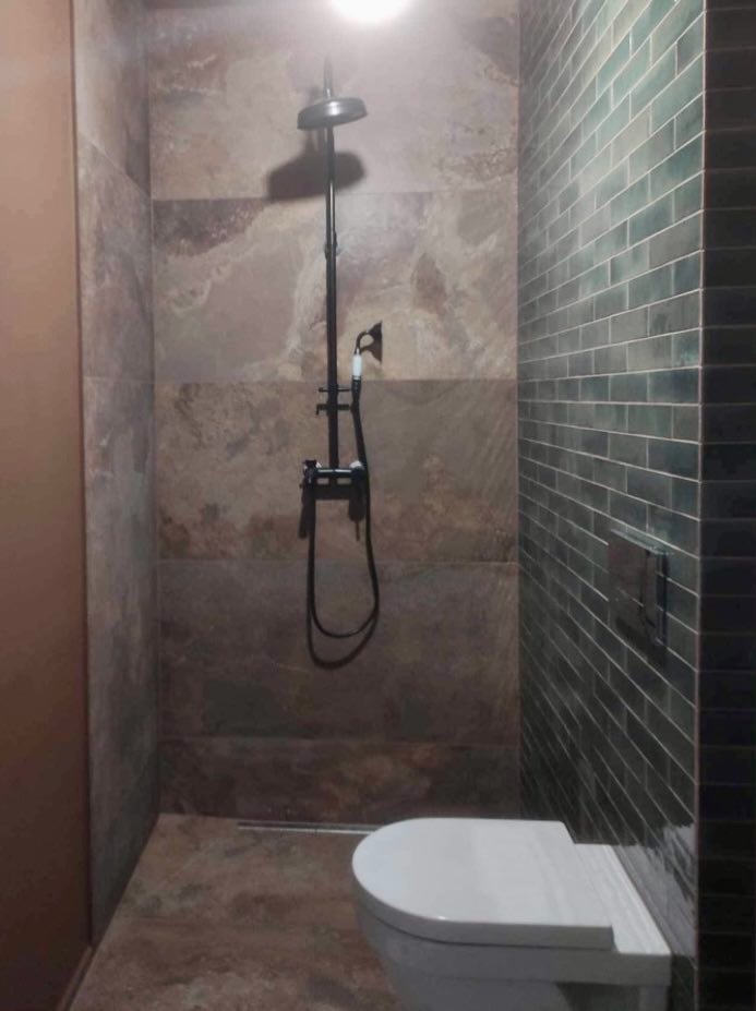 Wykończona łazienka z kabiną prysznicową typu walk-in, wyłożona płytkami imitującymi kamień na jednej ścianie i ciemnozielonymi, wąskimi płytkami na drugiej, z czarną armaturą i białą miską WC.