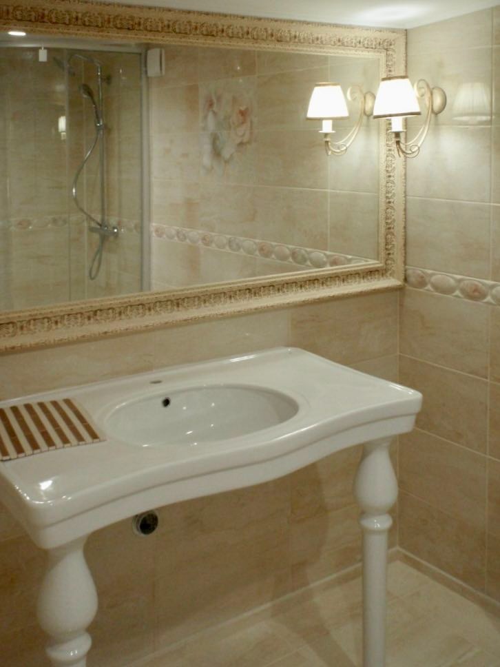 Jasna łazienka w Raniżowie z beżowymi płytkami, lustrem w ozdobnej ramie odbijającym kabinę prysznicową i umywalką na toczonych nogach z drewnianą kratką ociekową.