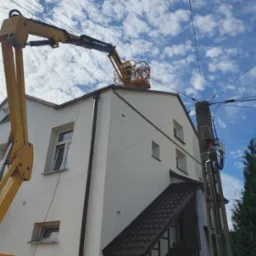 Pracownik w żółtym podnośniku koszowym na dachu dwupiętrowego budynku z białą elewacją, wykonujący prace na tle błękitnego nieba z obłokami. Widoczny słup energetyczny z przewodami.