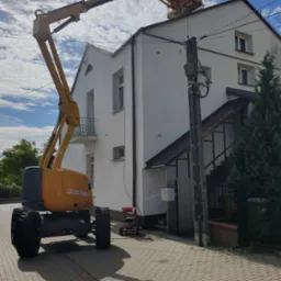 Pracownik na podnośniku koszowym maluje górną część elewacji dwupiętrowego domu z balkonem, widoczny także fragment chodnika z kostki brukowej i słup elektryczny.