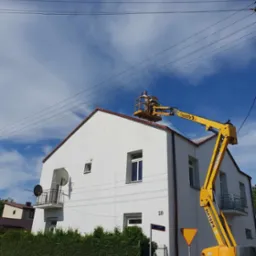 Pracownik na podnośniku Haulotte HA16PX, zabezpieczony w kasku, pracuje na dachu dwupiętrowego, białego domu z czerwonym dachem, na tle błękitnego nieba z chmurami.