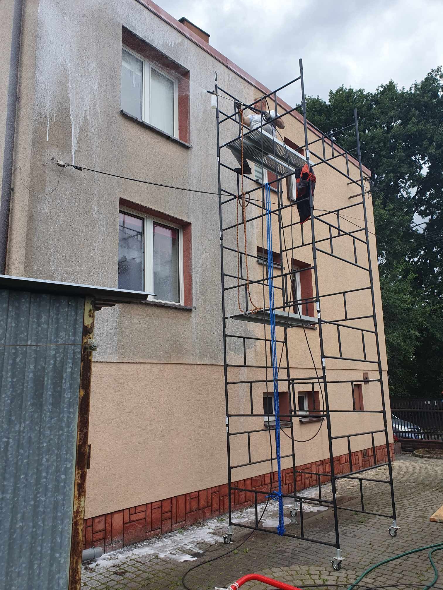 Pracownik na rusztowaniu czyści elewację budynku spryskując ją wodą pod ciśnieniem, widoczne zacieki na ścianie i mokra kostka brukowa.