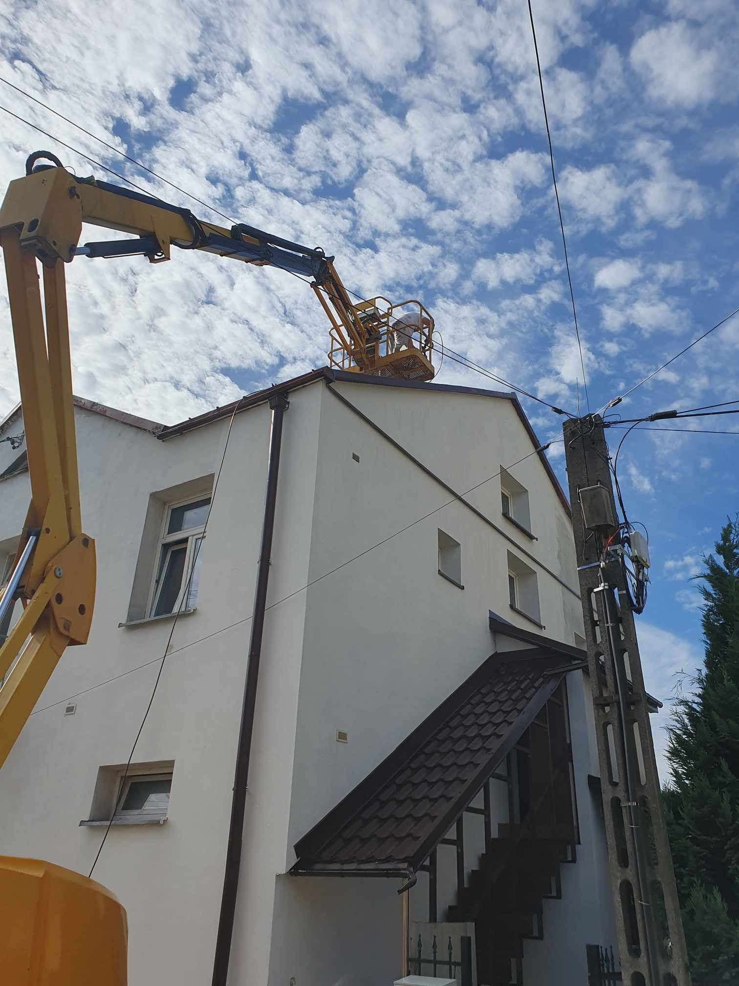Pracownik w żółtym podnośniku koszowym na dachu dwupiętrowego budynku z białą elewacją, wykonujący prace na tle błękitnego nieba z obłokami. Widoczny słup energetyczny z przewodami.