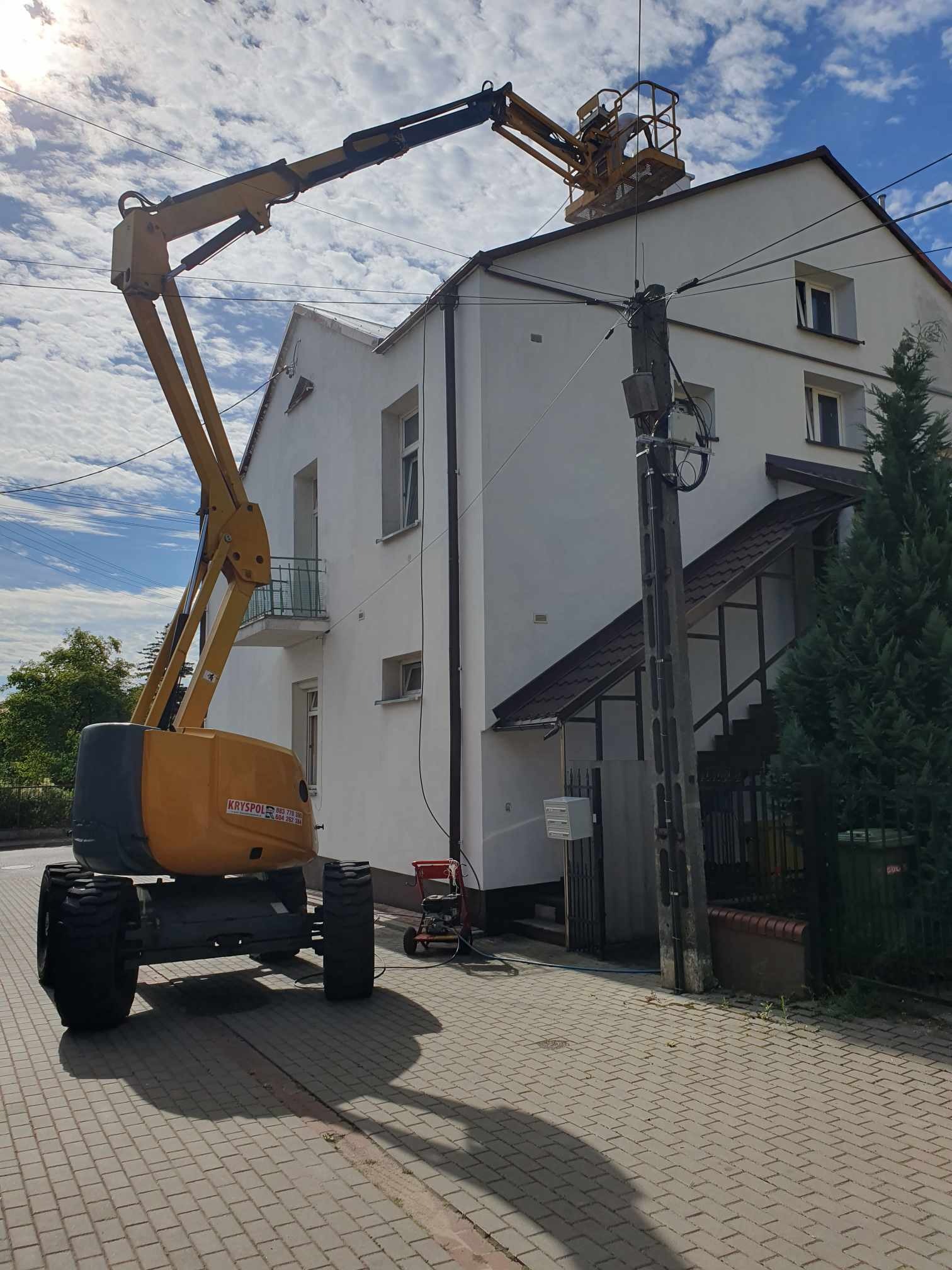 Pracownik na podnośniku koszowym maluje górną część elewacji dwupiętrowego domu z balkonem, widoczny także fragment chodnika z kostki brukowej i słup elektryczny.