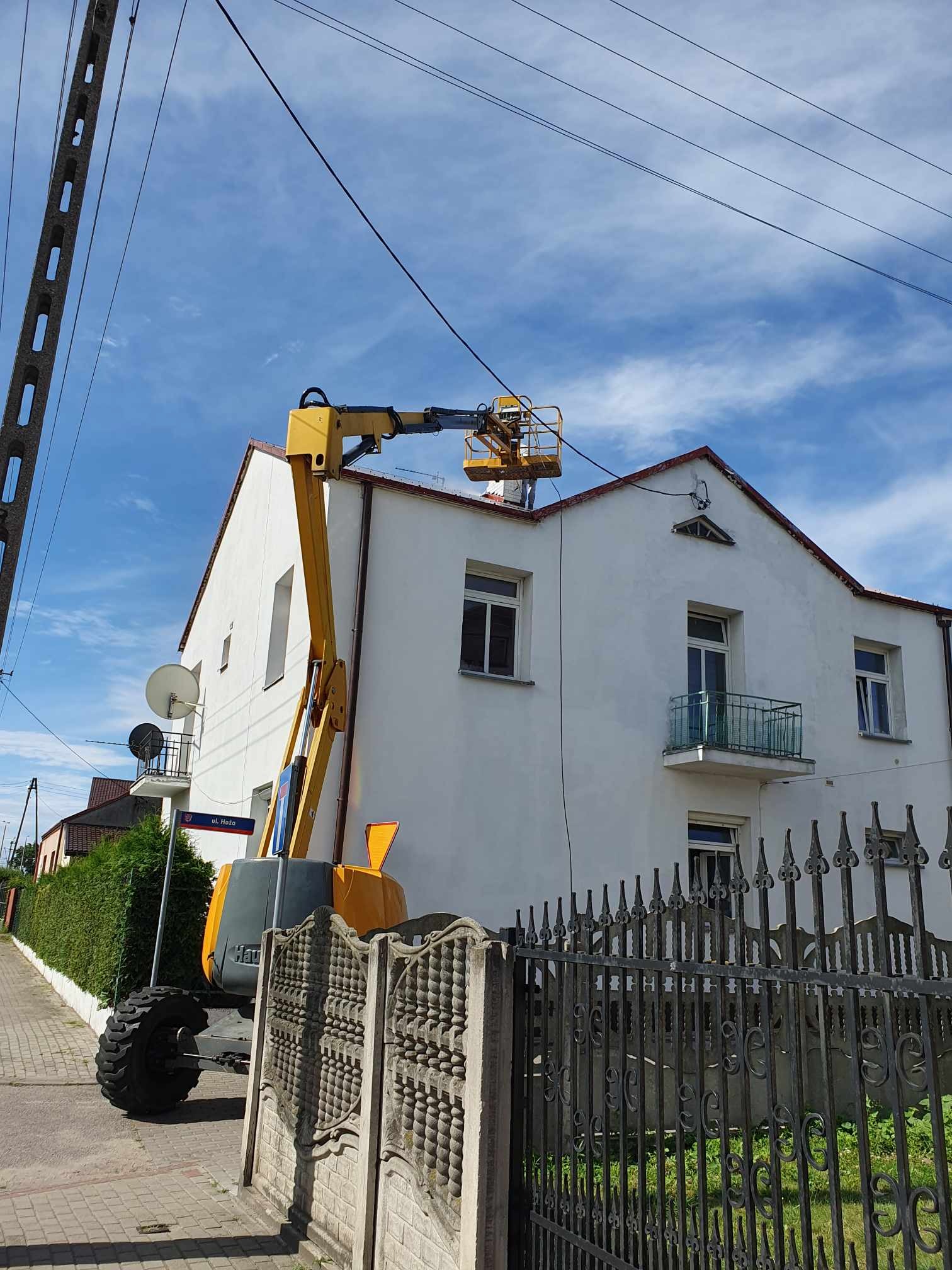 Żółty podnośnik koszowy na tle białej elewacji domu z czerwonym dachem, widoczne okna, balkon i ozdobne ogrodzenie, błękitne niebo w tle.