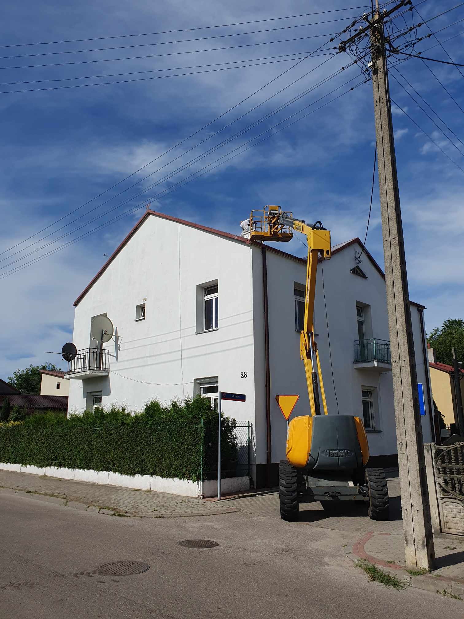 Pracownik w podnośniku koszowym na wysokości, wykonujący prace na dachu dwupiętrowego, białego budynku z balkonami, widok z poziomu ulicy w słoneczny dzień.