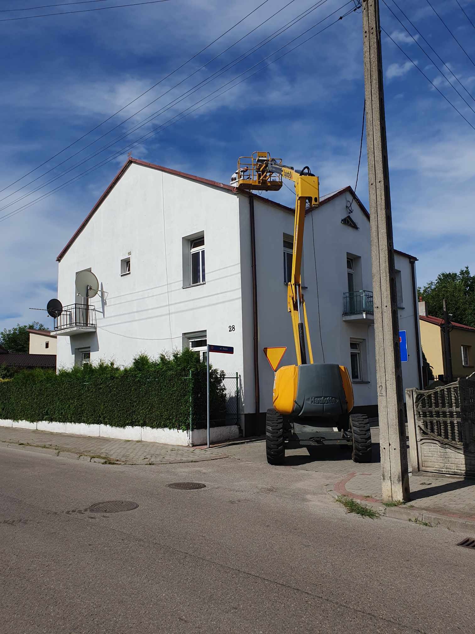 Żółto-szara platforma podnośnikowa na tle dwupiętrowego, białego domu z brązowym dachem, podczas prac na wysokości dachu.