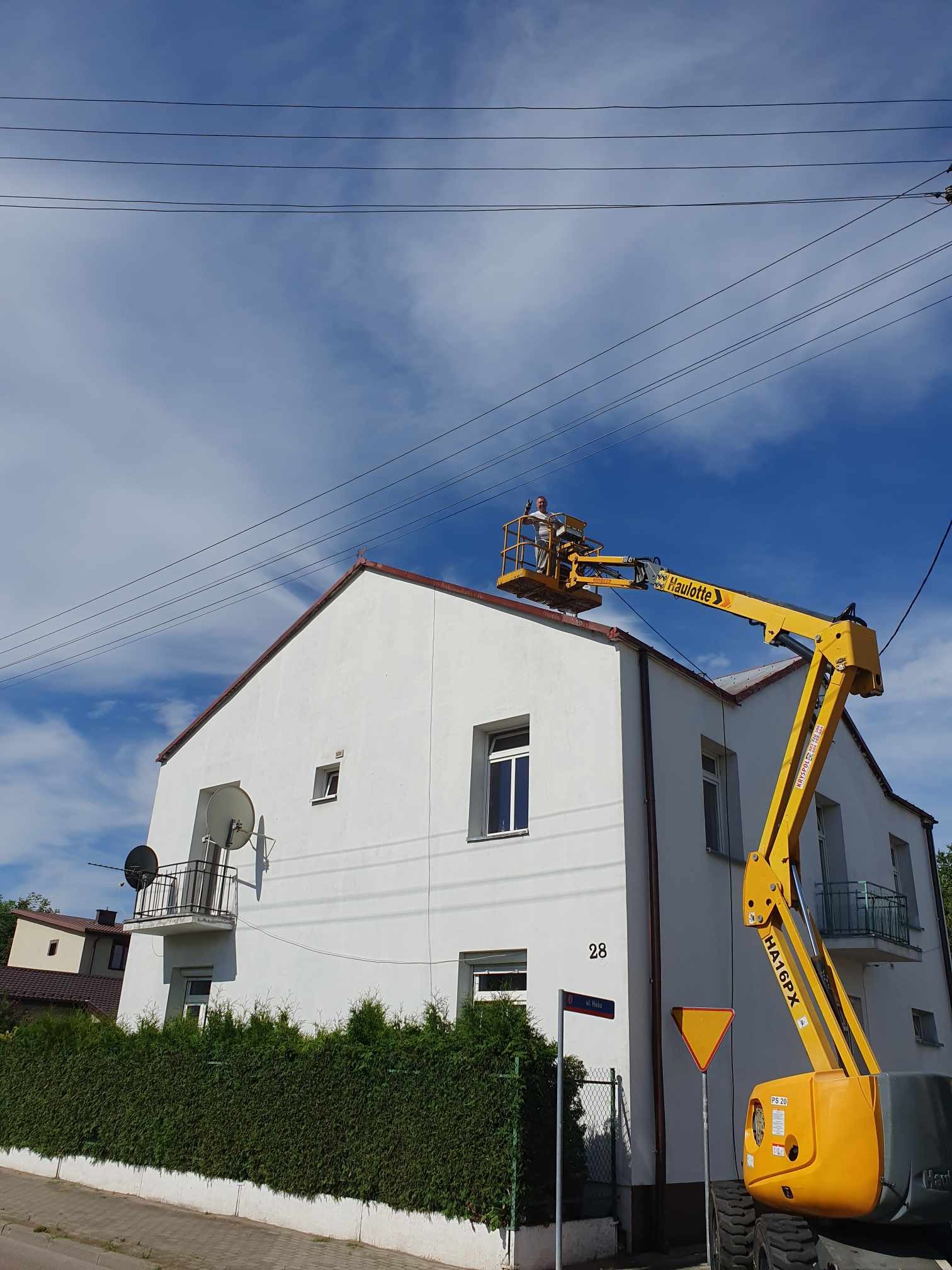 Pracownik na podnośniku Haulotte HA16PX, zabezpieczony w kasku, pracuje na dachu dwupiętrowego, białego domu z czerwonym dachem, na tle błękitnego nieba z chmurami.