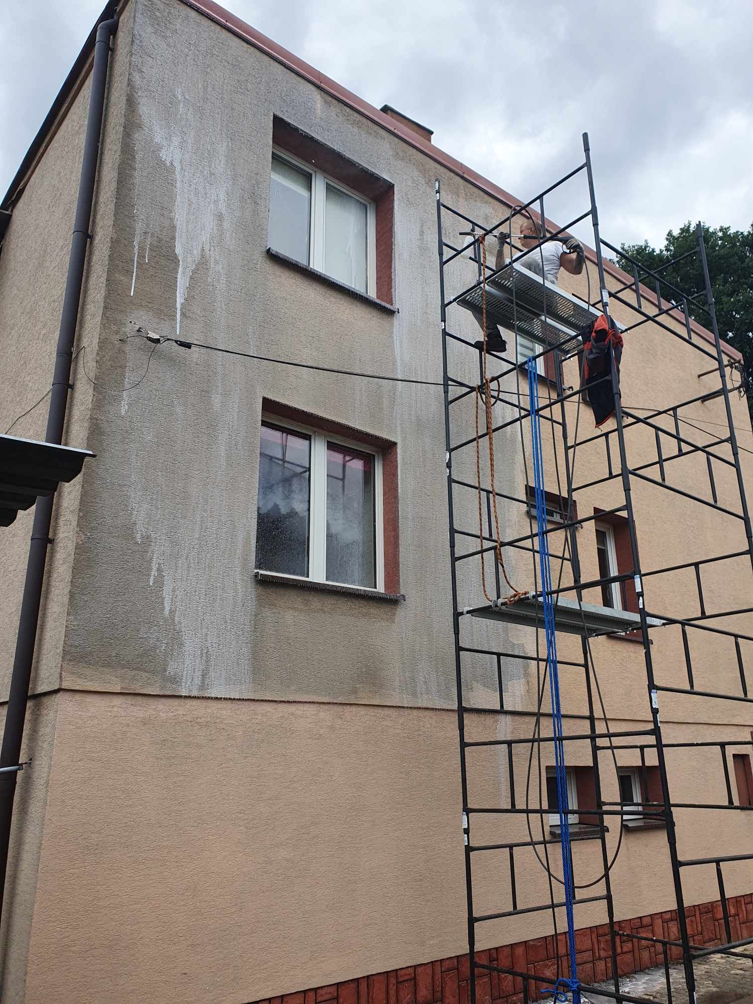 Pracownik na rusztowaniu, podczas mycia elewacji budynku, widoczne zacieki wody na szarej części fasady, a także pomarańczowa część elewacji i okna.