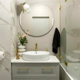 Nowoczesna łazienka z szarą szafką pod umywalkę, okrągłym lustrem i złotymi akcentami, takimi jak bateria, uchwyty i rama kabiny prysznicowej, oświetlona lampą kulistą.