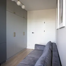 Szara zabudowa szaf wnękowych z złotymi uchwytami, oświetlenie sufitowe z trzema kulistymi lampami, szara sofa i minimalistyczne obrazy w białych ramach.