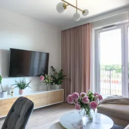 Jasne wnętrze salonu z drewnianą szafką RTV, dekoracjami roślinnymi, marmurowymi stolikami kawowymi ze złotymi nogami oraz modernistyczną lampą sufitową z białymi kulami