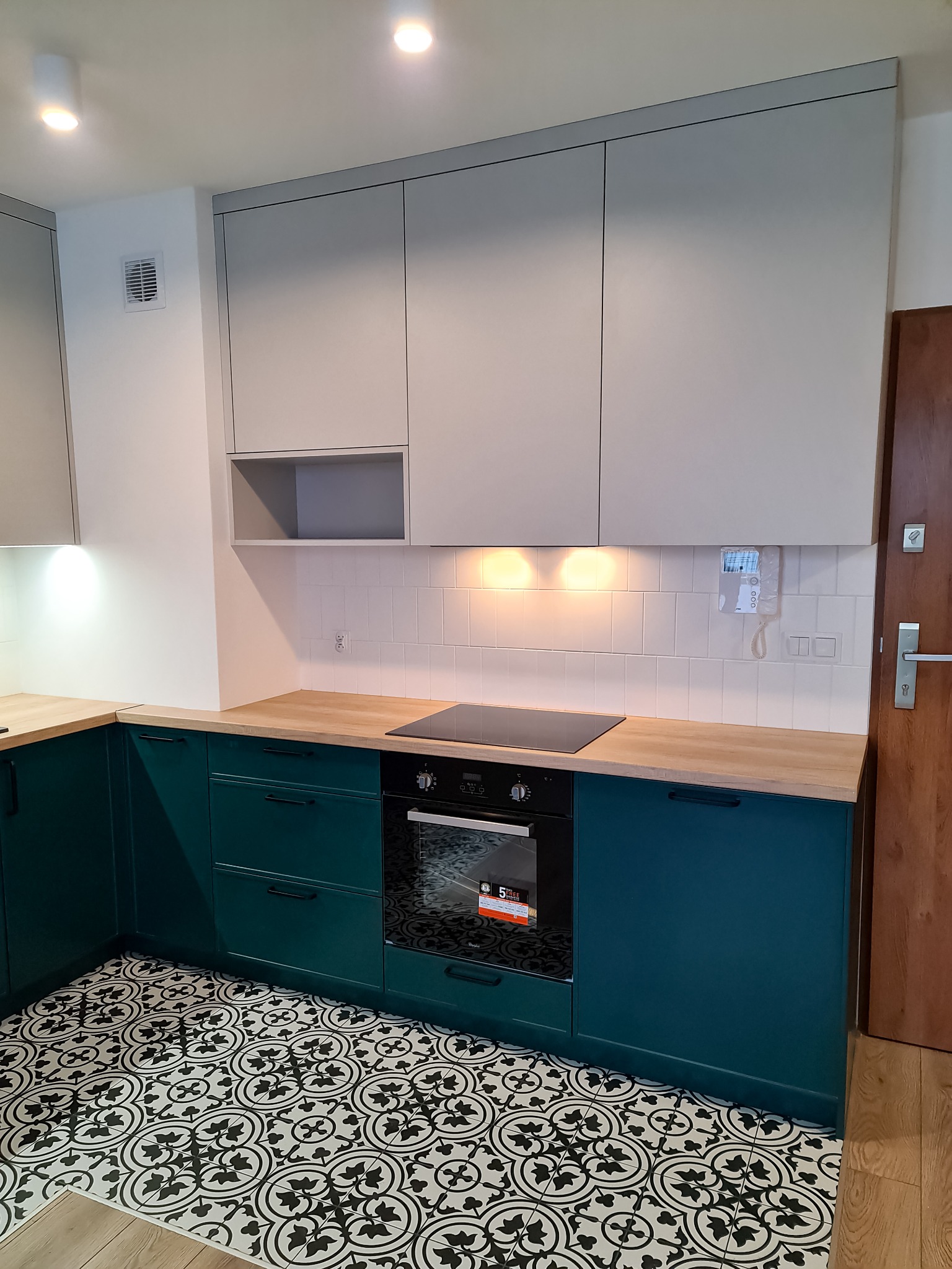 Nowoczesna kuchnia z szarymi szafkami górnymi i butelkowozielonymi dolnymi, drewnianym blatem oraz czarno-białą wzorzystą podłogą. Widoczny piekarnik z płytą indukcyjną i białe płytki na ścianie.