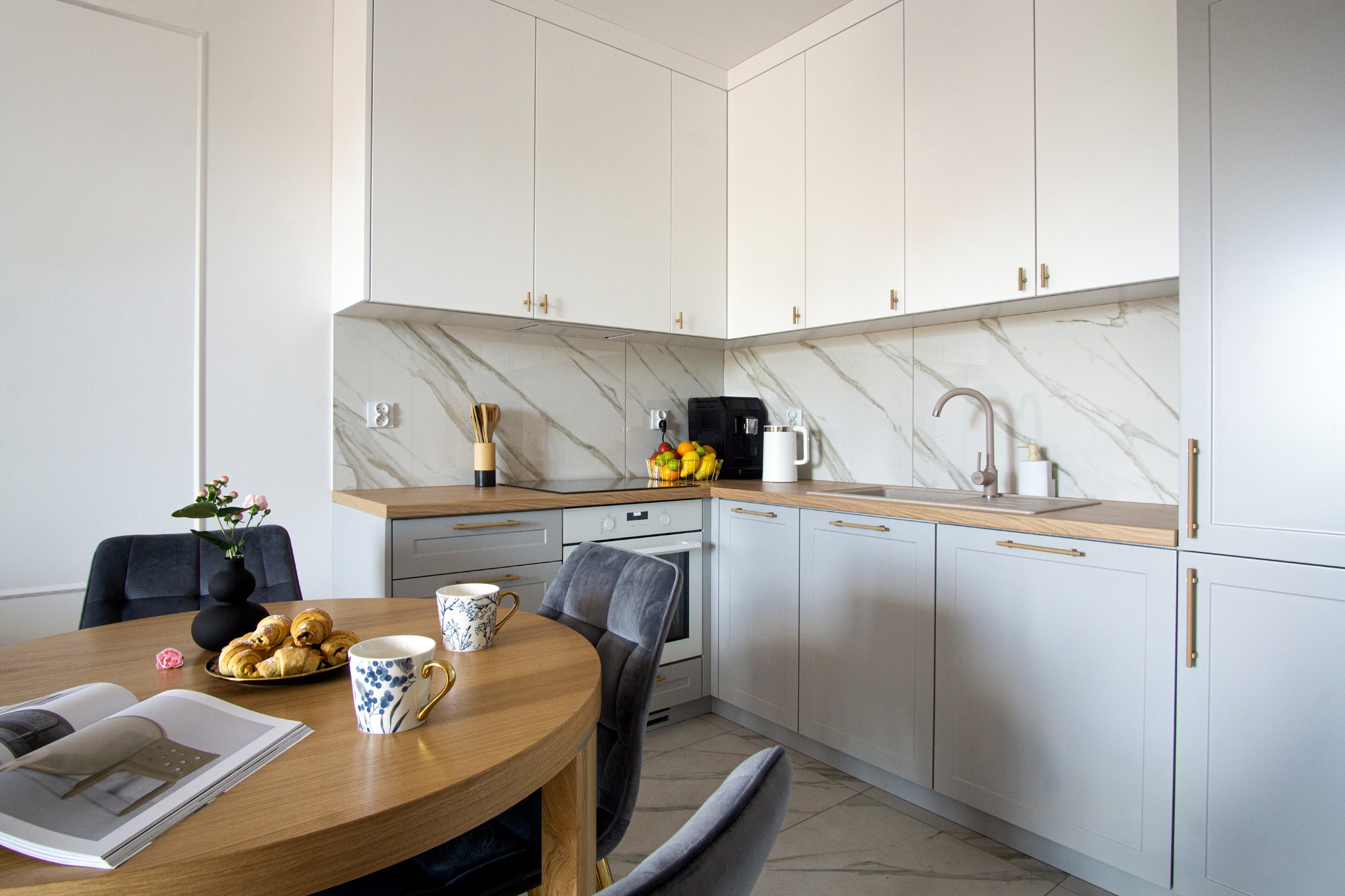 Jasna kuchnia z drewnianym blatem, marmurową ścianą i okrągłym stołem z rogalikami i filiżankami, nowoczesny design.