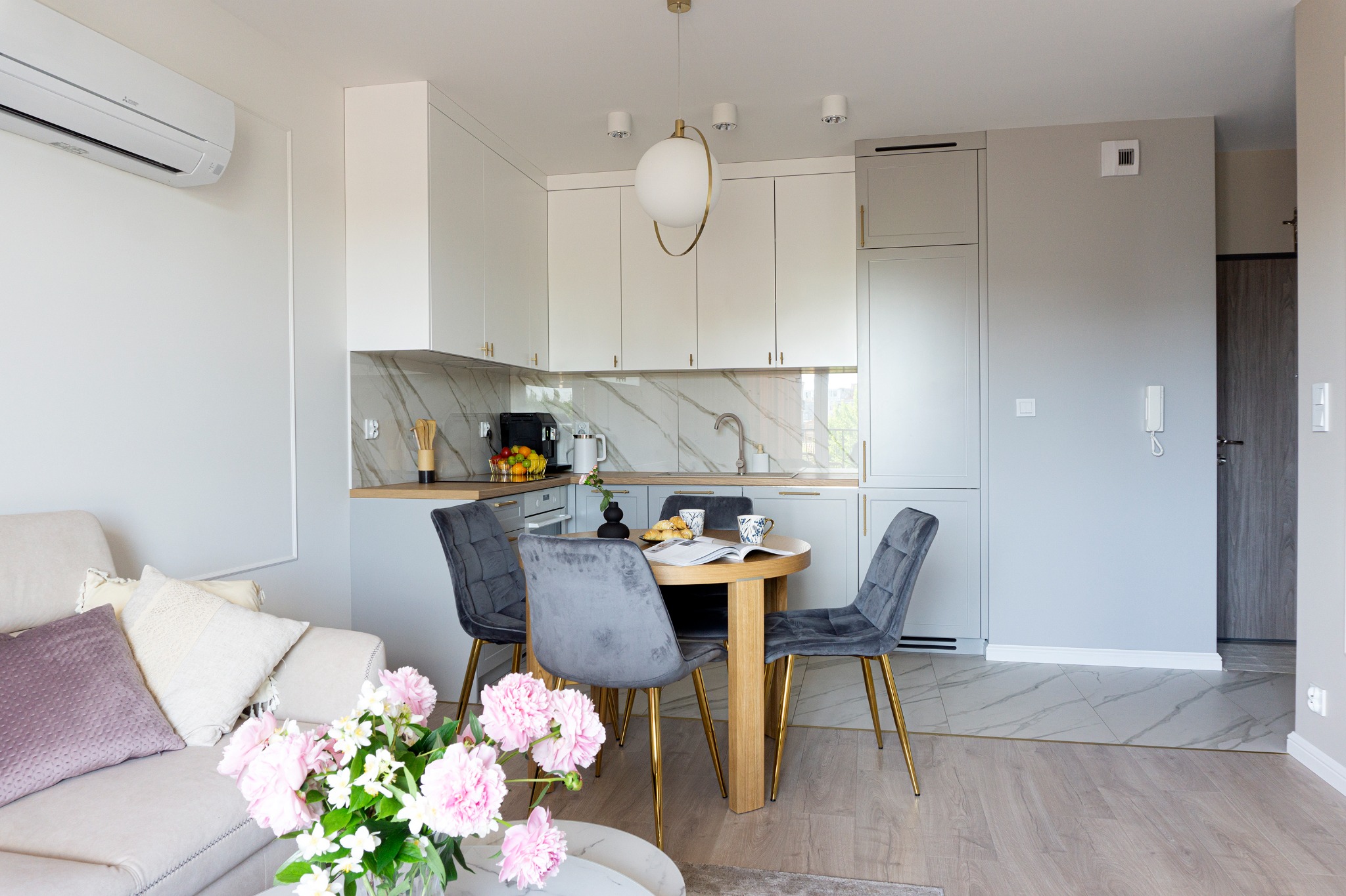 Jasna kuchnia w stylu skandynawskim z marmurowym backsplashiem, okrągłym drewnianym stołem i szarymi krzesłami na złotych nogach, całość doświetlona kulistą lampą.
