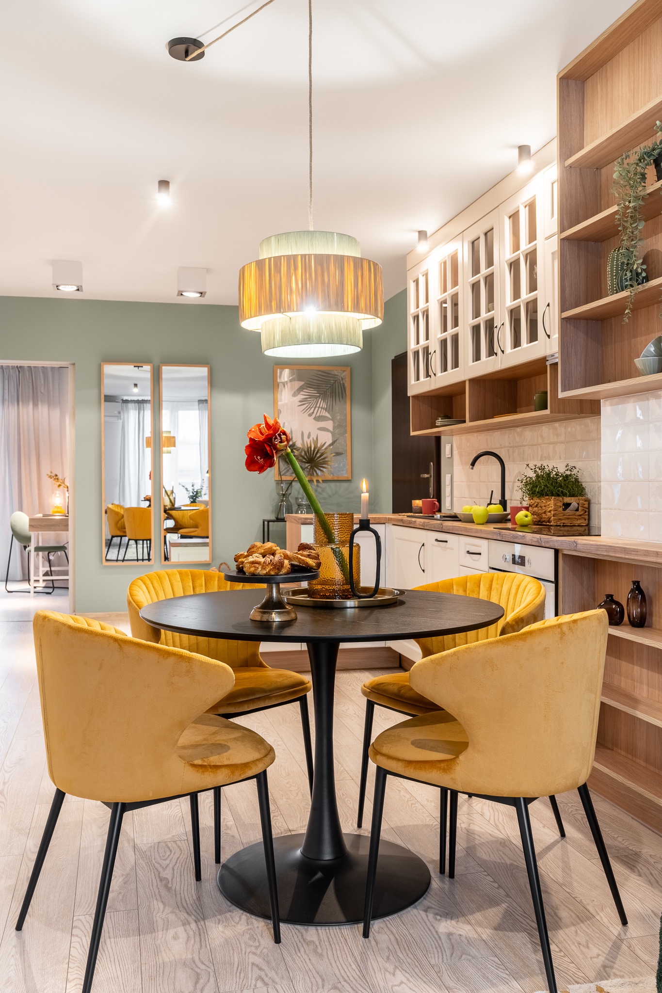 Stylowa aranżacja jadalni z okrągłym stołem, złotymi krzesłami i kwiatem amarylisa w wazonie. W tle kuchnia z drewnianymi półkami i lustra.