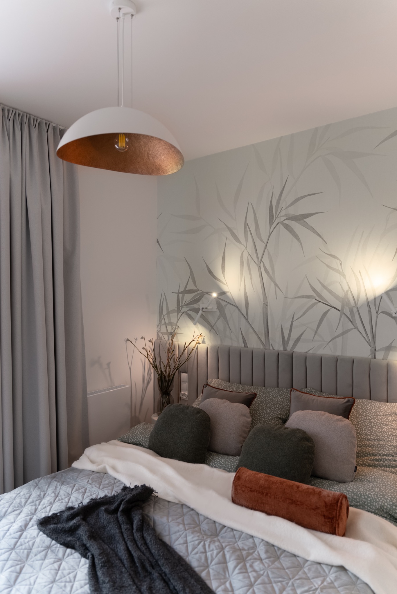 Stylowa sypialnia z łóżkiem, szarą tapetą z motywem roślinnym i lampą wiszącą. Widoczne poduszki dekoracyjne i pled na łóżku. Przytulne wnętrze.