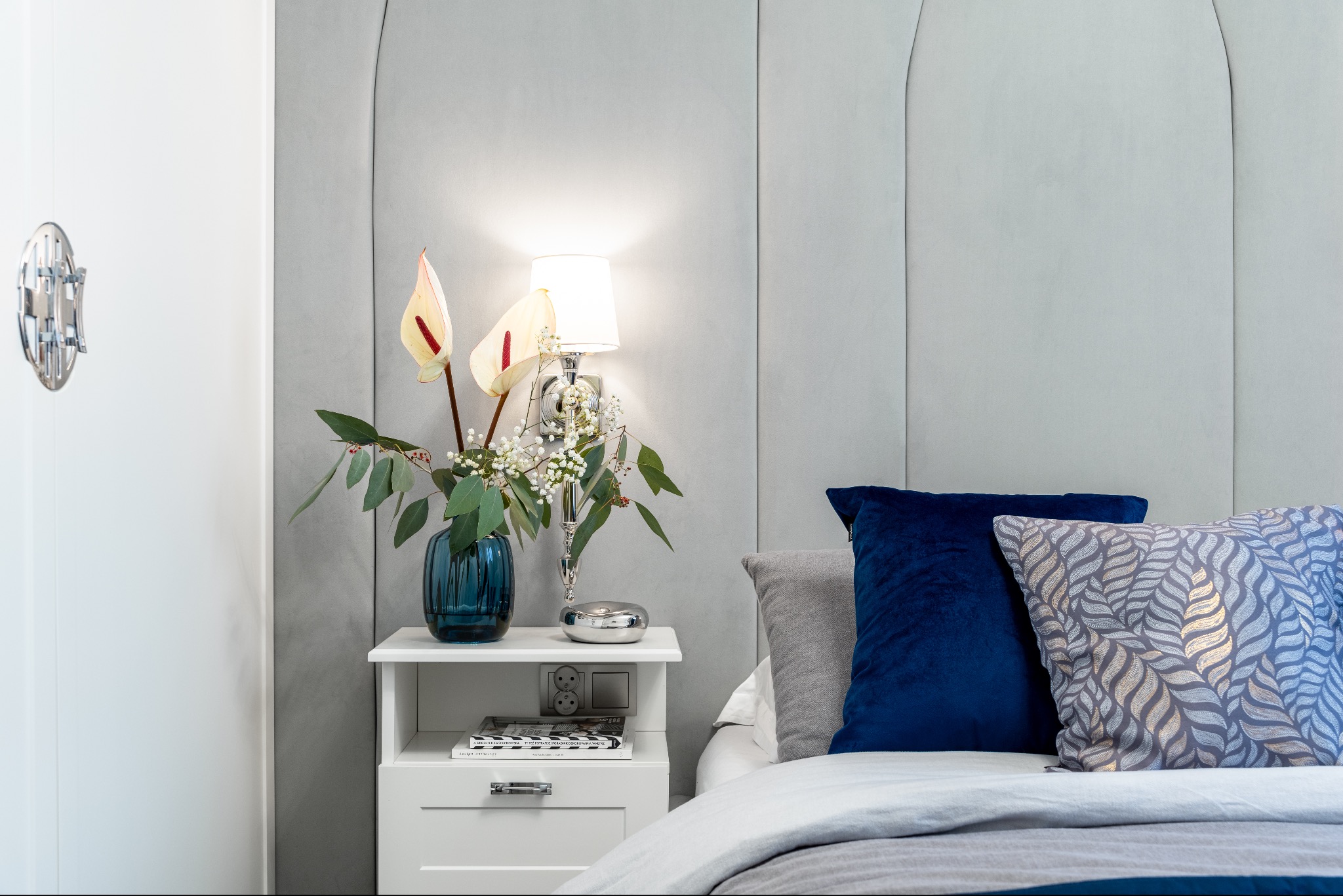 Aranżacja sypialni: nocna szafka z kwiatami anthurium, lampą i poduszkami na łóżku. Minimalistyczny design z szarym panelem ściennym i białymi meblami.