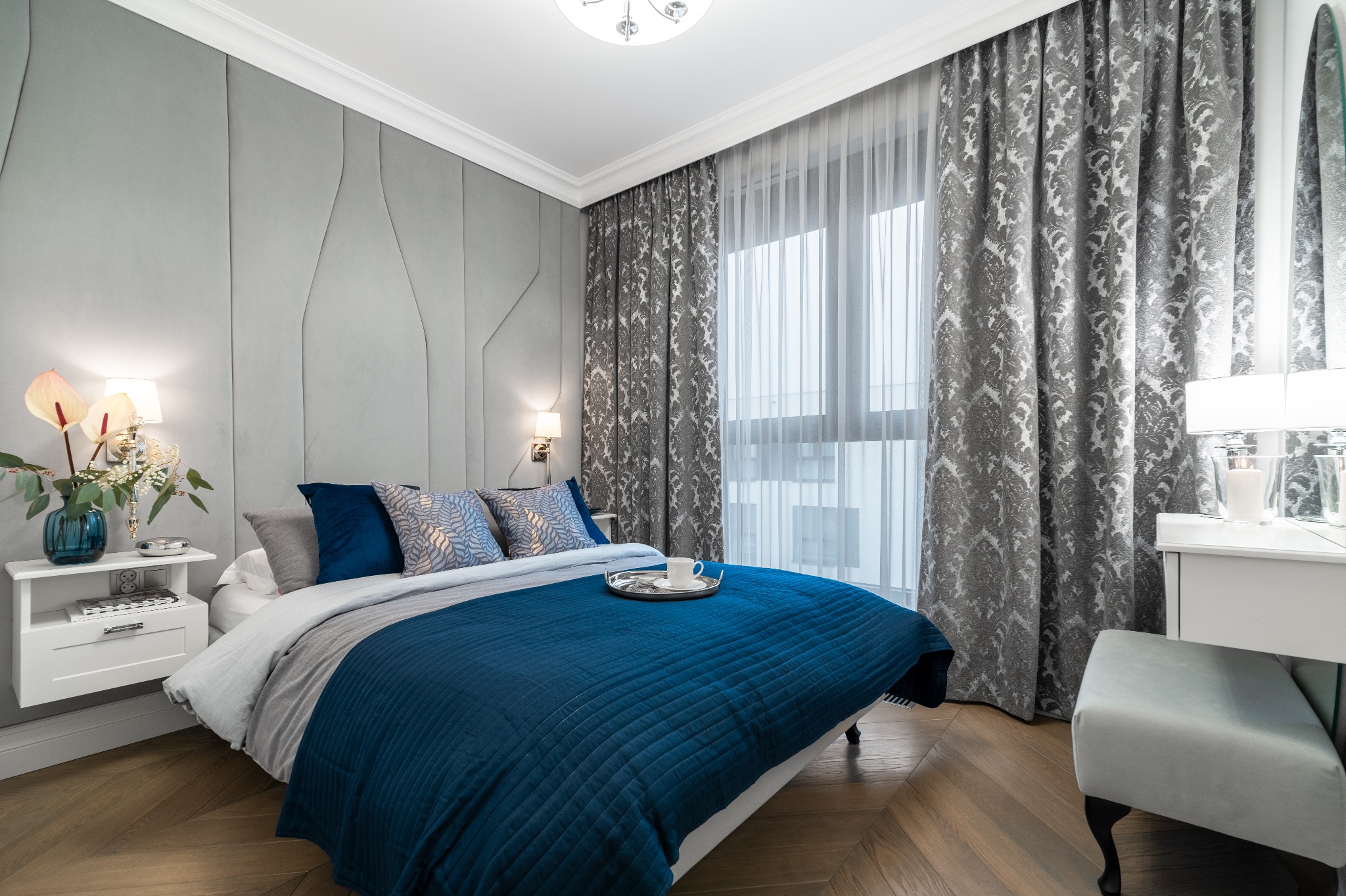 Stylowa sypialnia z łóżkiem w niebieskiej pościeli, szarą tapetą z geometrycznym wzorem i drewnianą podłogą w jodełkę. Na łóżku taca z filiżanką.