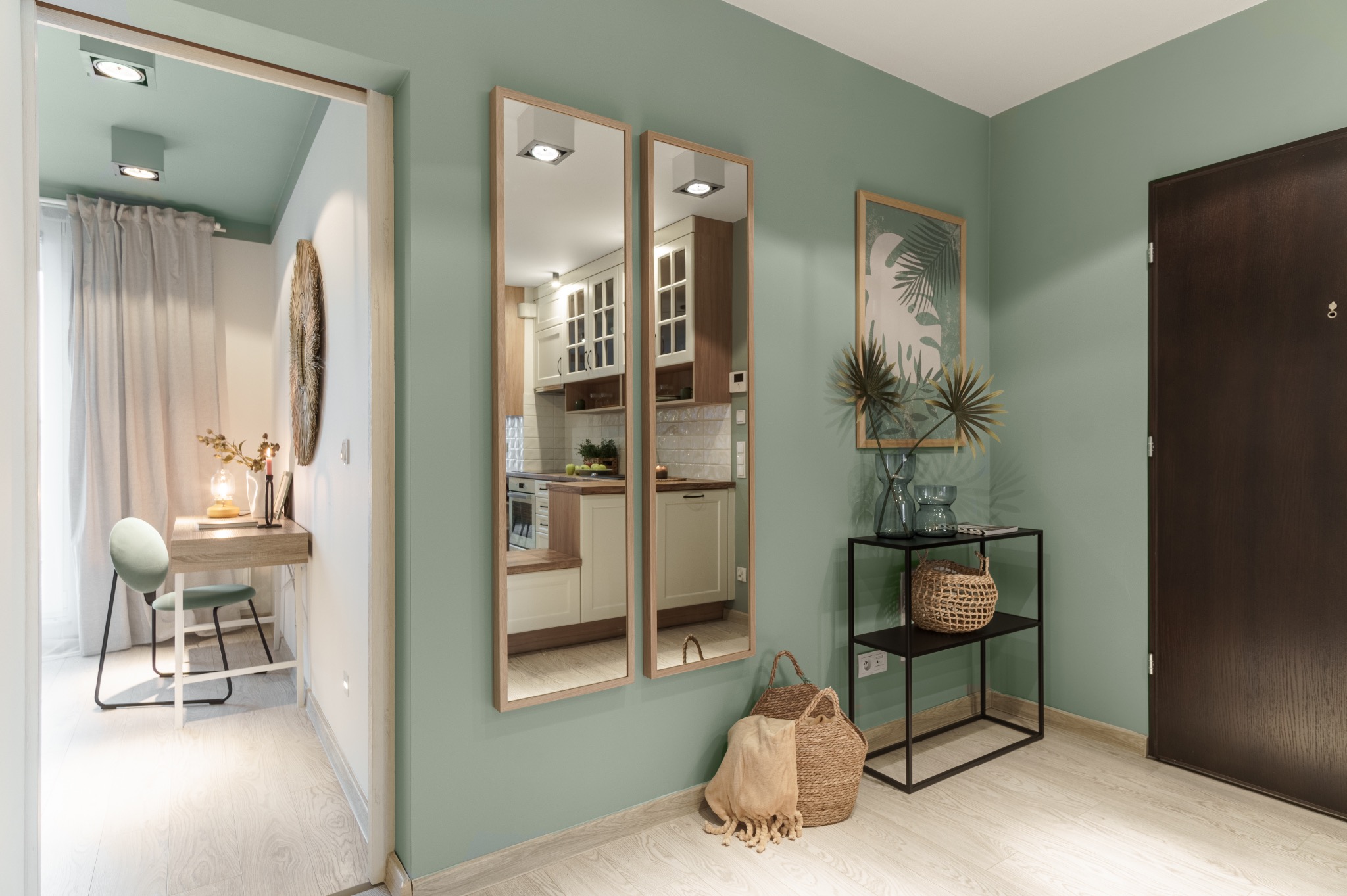 Stylowy przedpokój w odcieniach zieleni z lustrami odbijającymi wnętrze kuchni, drewnianą podłogą i minimalistycznymi dodatkami. Widok na biurko w pokoju obok.