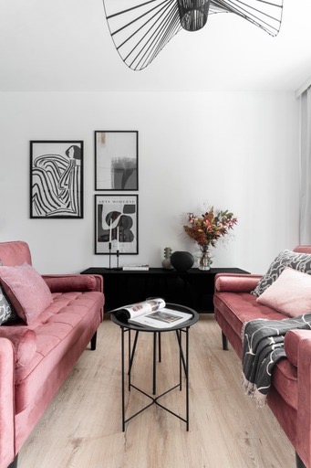 Stylowy salon z dwoma różowymi sofami, czarnym stolikiem kawowym i komodą. Na ścianie wiszą trzy grafiki, a całość dopełnia nowoczesna lampa.