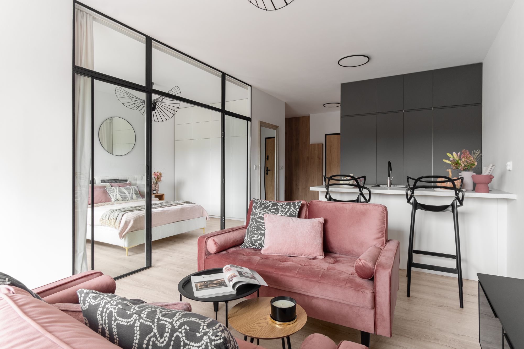 Jasne wnętrze mieszkania z różową sofą, czarnymi stolikami kawowymi i przeszkloną ścianą oddzielającą sypialnię. Nowoczesny design z elementami retro.