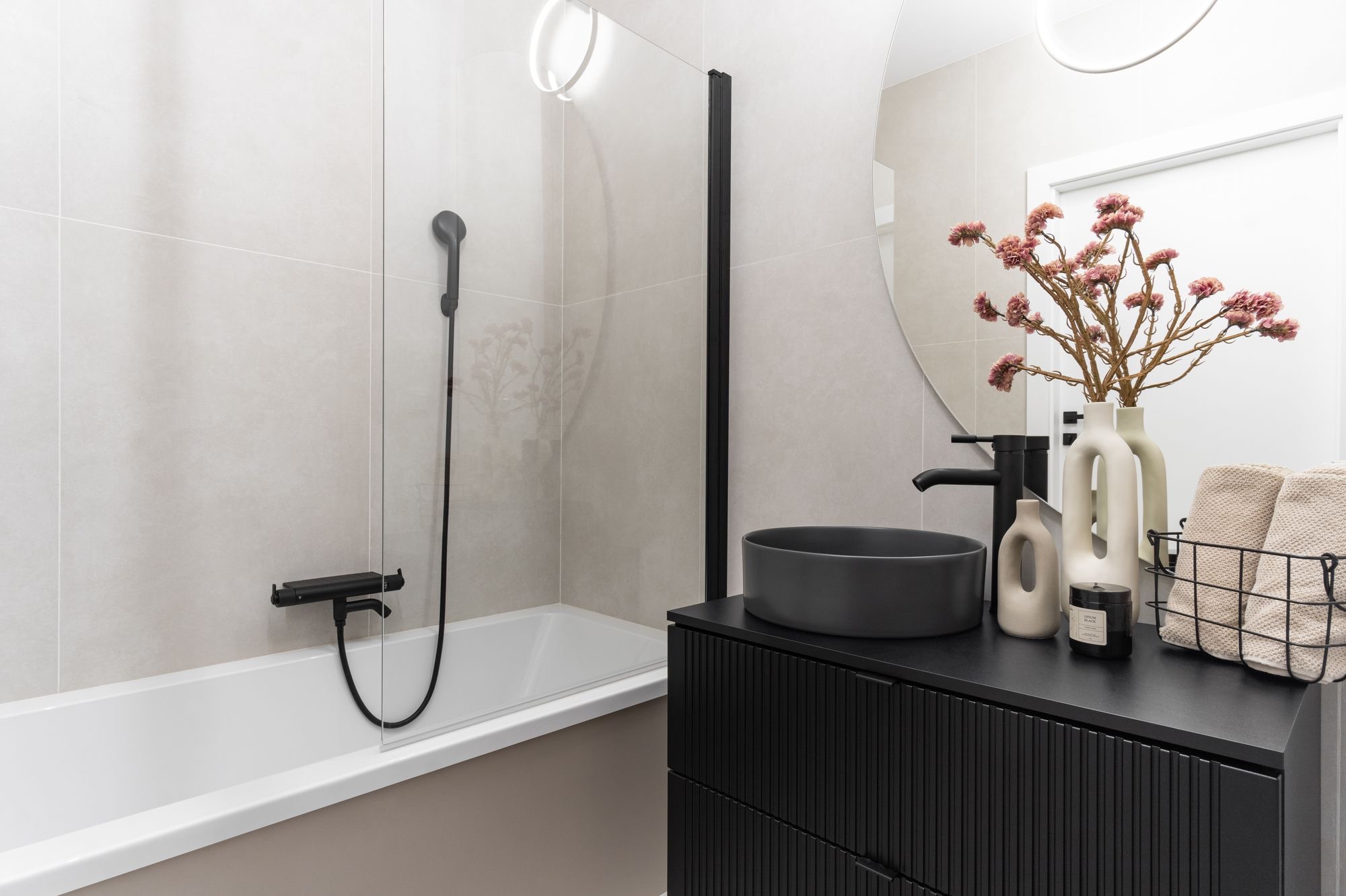 Nowoczesna łazienka z czarną armaturą i umywalką nablatową. Minimalistyczny design, beżowe płytki, okrągłe lustro i dekoracyjne akcenty.